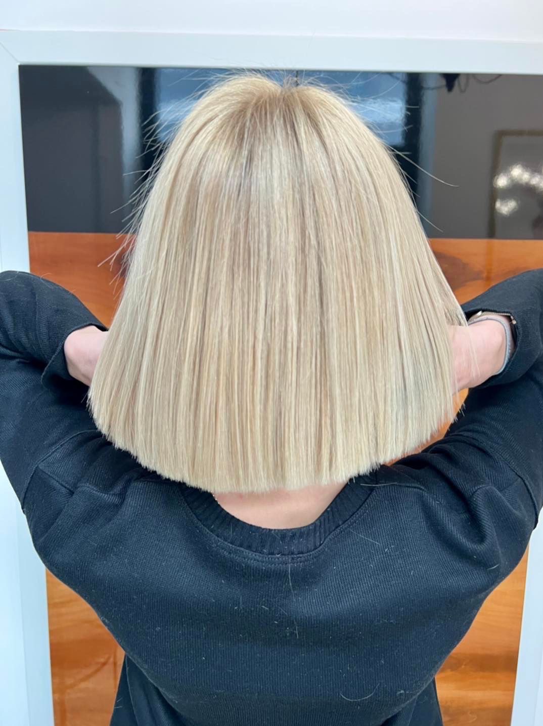 Blond włosy ścięte na prosto, widok z tyłu, kobieta w czarnej bluzie trzyma ręce za głową, na tle drewnianej ściany.