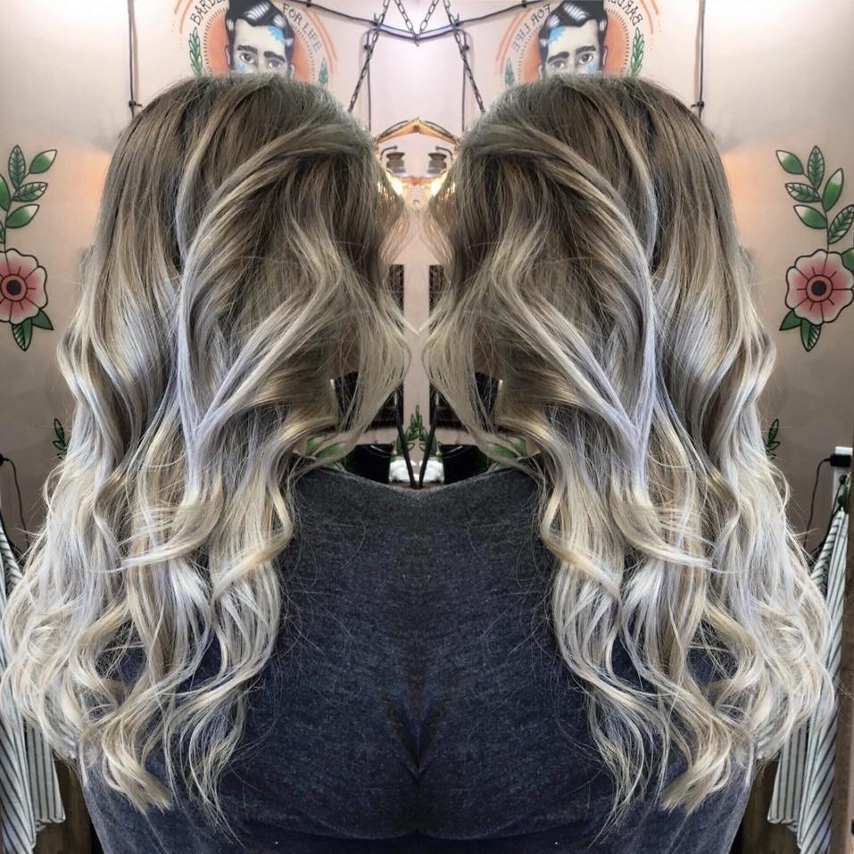 Długie, falowane włosy z ombre w odcieniach blondu i szarości, widoczne od tyłu na tle ściany z naklejkami w stylu retro.