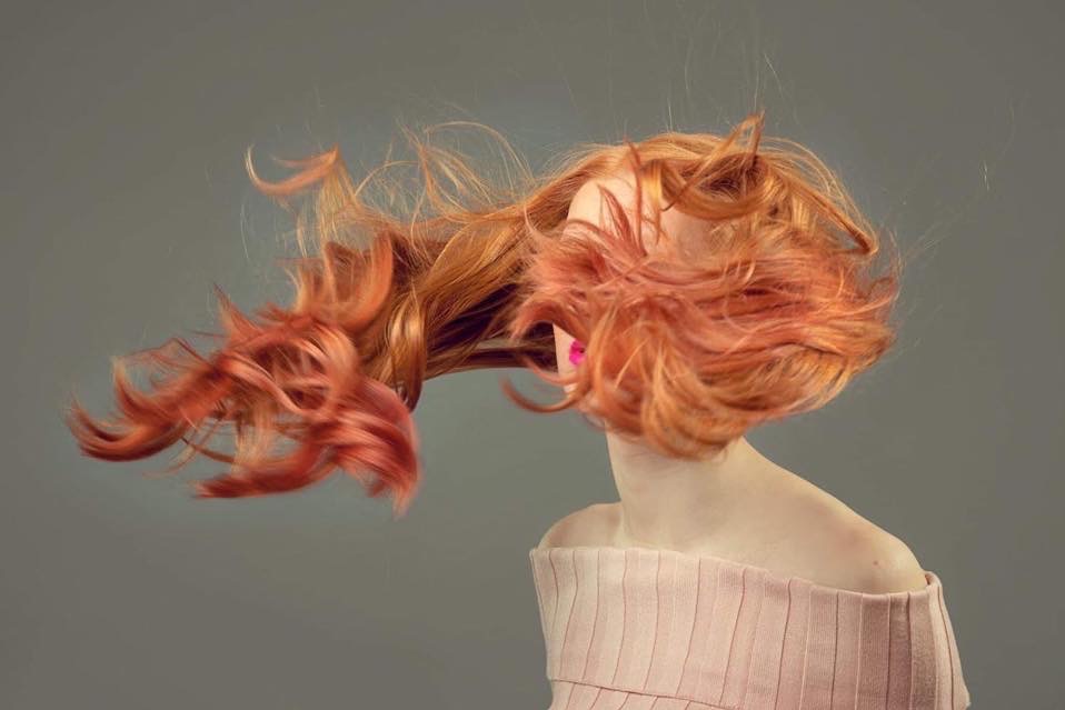 Dynamiczne ujęcie rudych włosów w ruchu, zarys profilu kobiety z jaskrawą szminką na tle szarego tła.