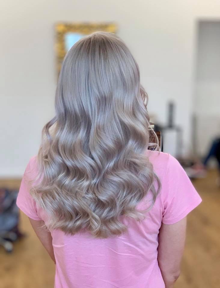 Tył głowy kobiety z długimi, falowanymi włosami w chłodnym odcieniu blondu, widoczne odbicie lustra w tle.