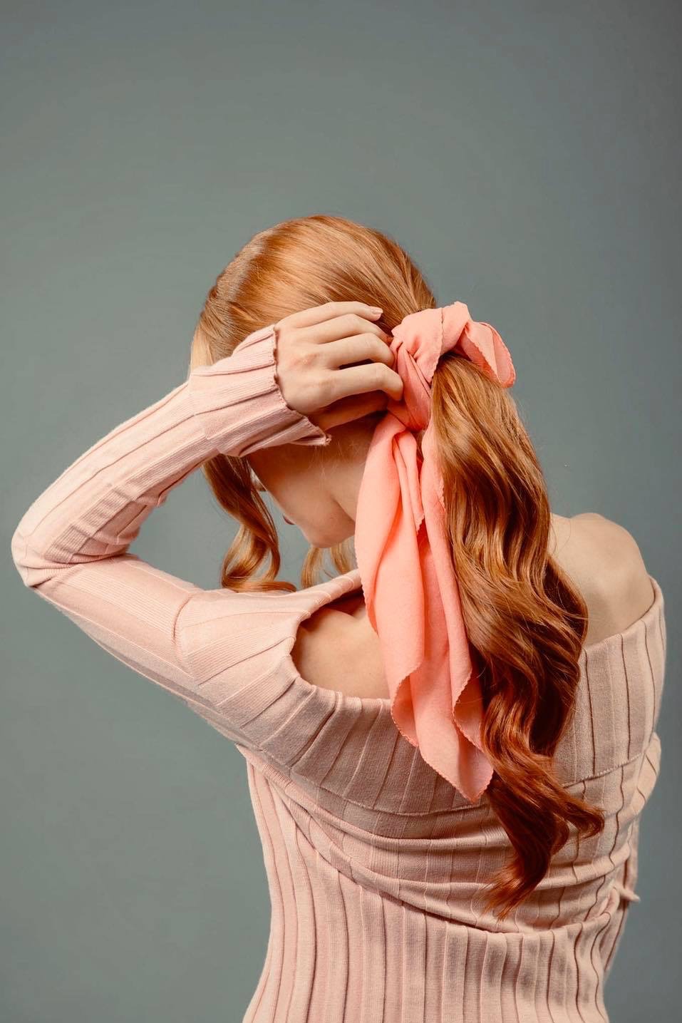 Rudowłosa kobieta z upiętymi włosami, związana różową apaszką, prezentowana od tyłu, ubrana w dopasowany sweter z odkrytymi ramionami na jednolitym szarym tle.