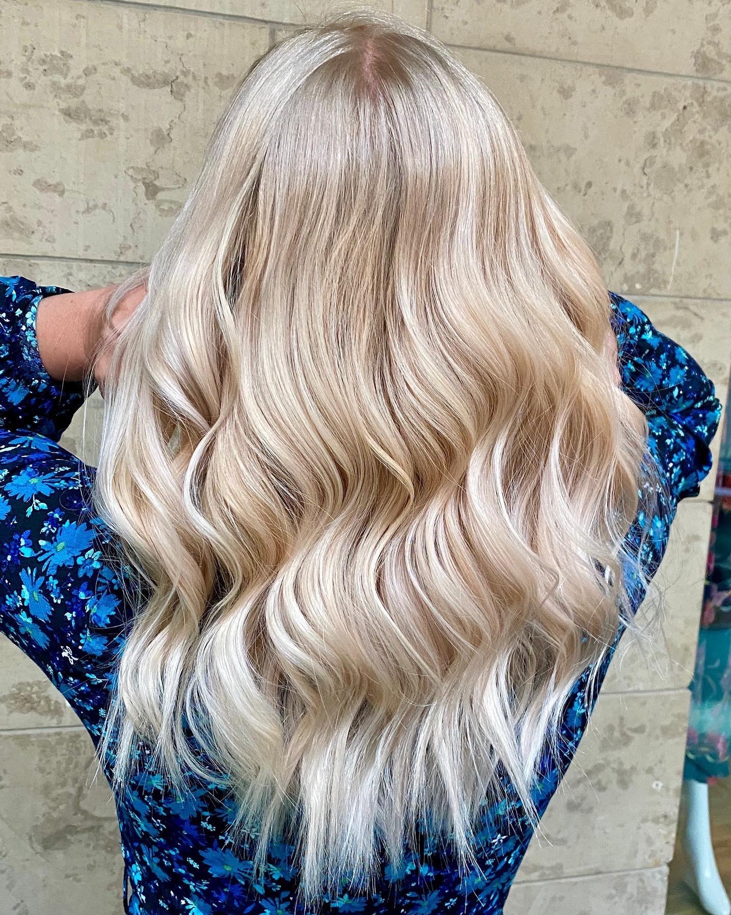 Tył głowy kobiety z długimi, falowanymi, platynowymi włosami z subtelnymi refleksami, ubrana w sukienkę w niebieskie kwiaty, na tle ściany o teksturze marmuru.