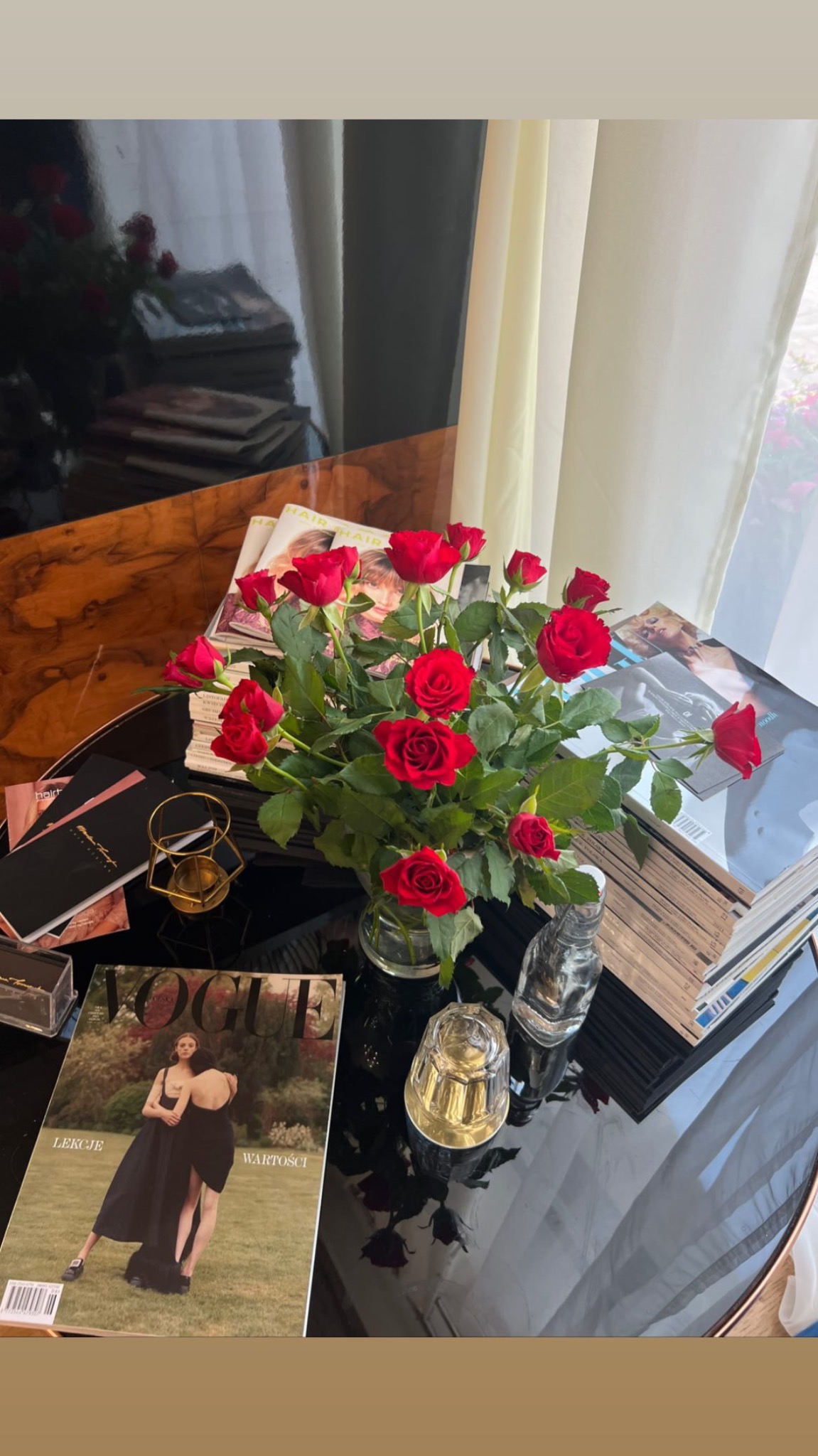 Stół z czarnym blatem, na którym leżą magazyny modowe, wazon z czerwonymi różami i szklane dekoracje, obok okna z jasnymi zasłonami.