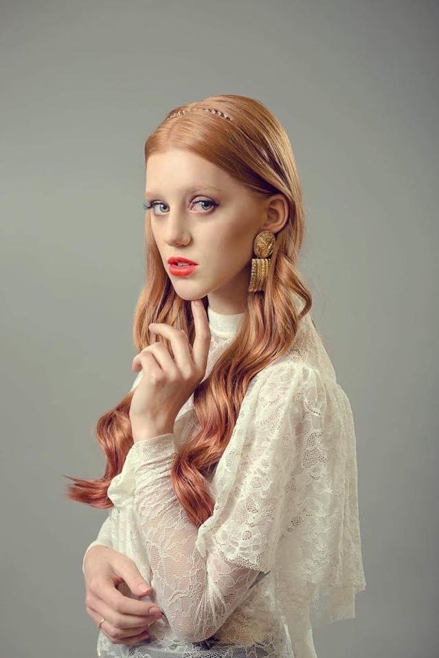 Portret kobiety z rudymi włosami w stylu retro, z delikatnym makijażem i dużymi złotymi kolczykami, pozującej na szarym tle.