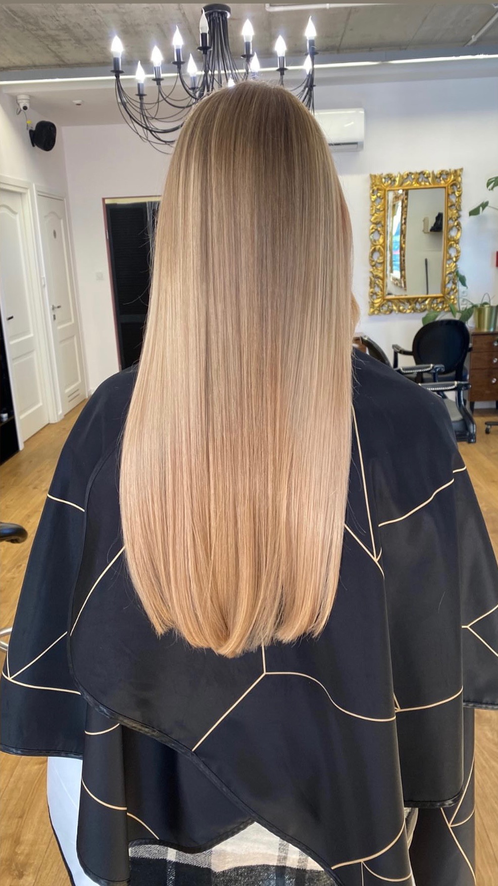Długie, proste włosy w odcieniach blondu, widok z tyłu, nałożona peleryna fryzjerska z geometrycznym wzorem w salonie z lustrem w złotej ramie i żyrandolem.