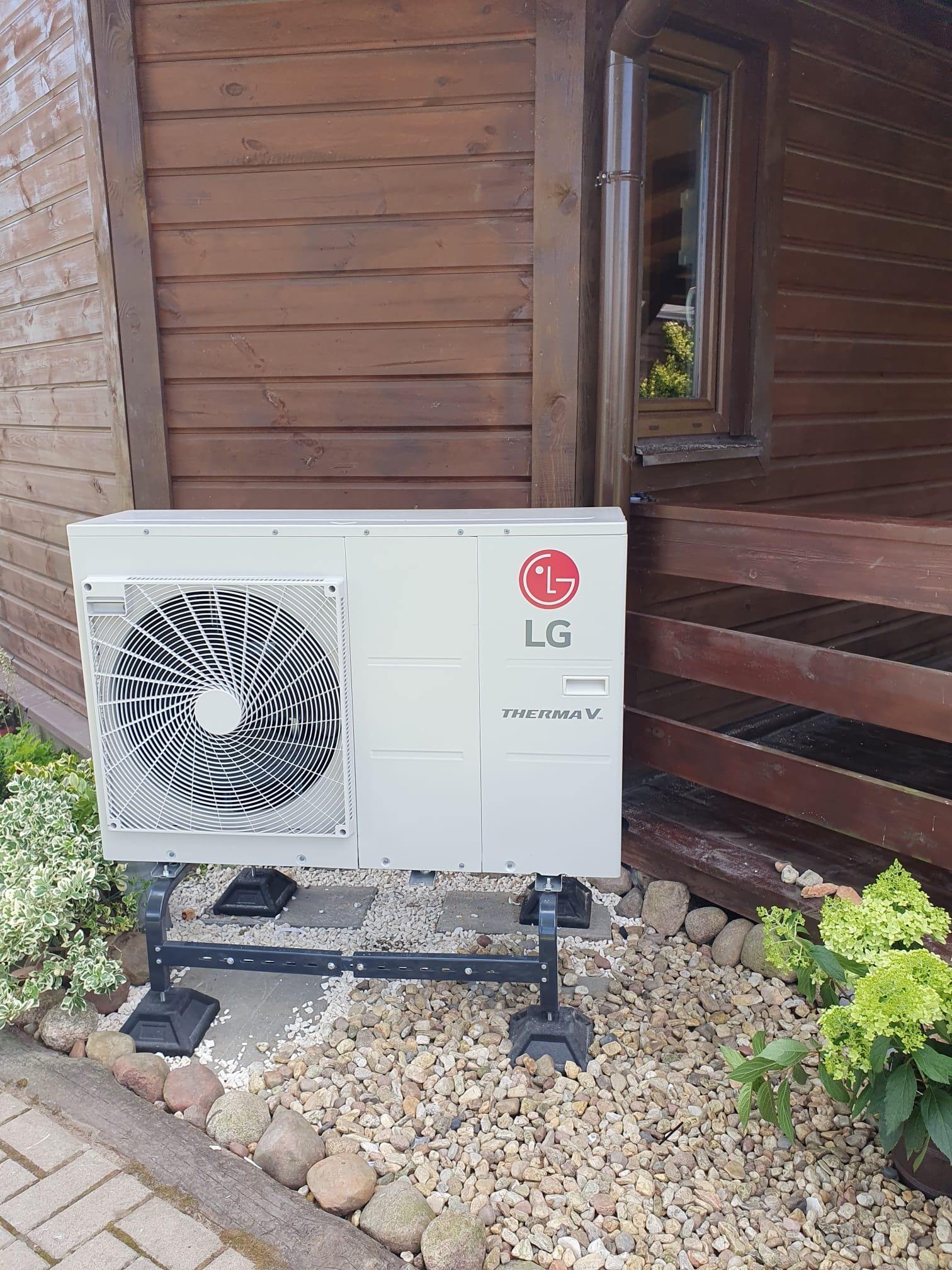Biała pompa ciepła LG Therma V zamontowana na czarnym stelażu przed drewnianym domem z jasnymi kamykami i zielenią wokół.