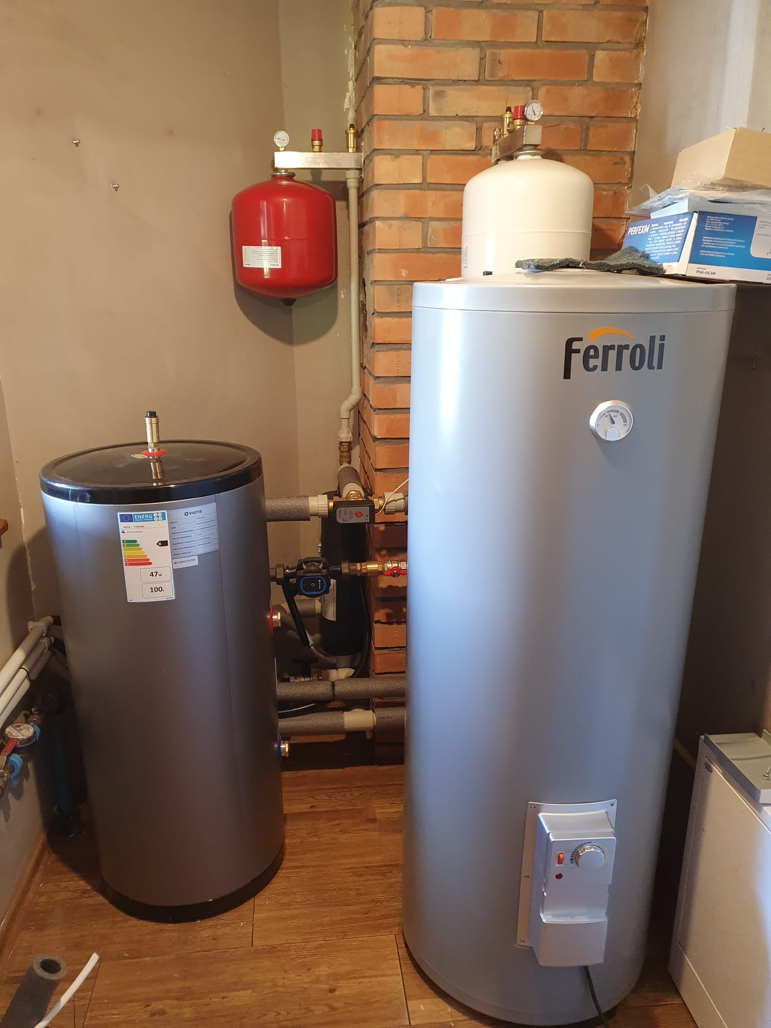 Dwa zbiorniki akumulacyjne ciepła różnych marek (Ferroli i Viotis) w kotłowni, z widocznymi rurami, zaworami i manometrami, na tle ściany z cegły i pomalowanej ściany.