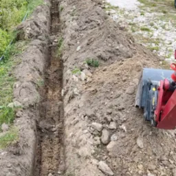 Długi, wąski wykop w ziemi, wykonany blisko domu jednorodzinnego, z widocznym fragmentem koparki z boku.