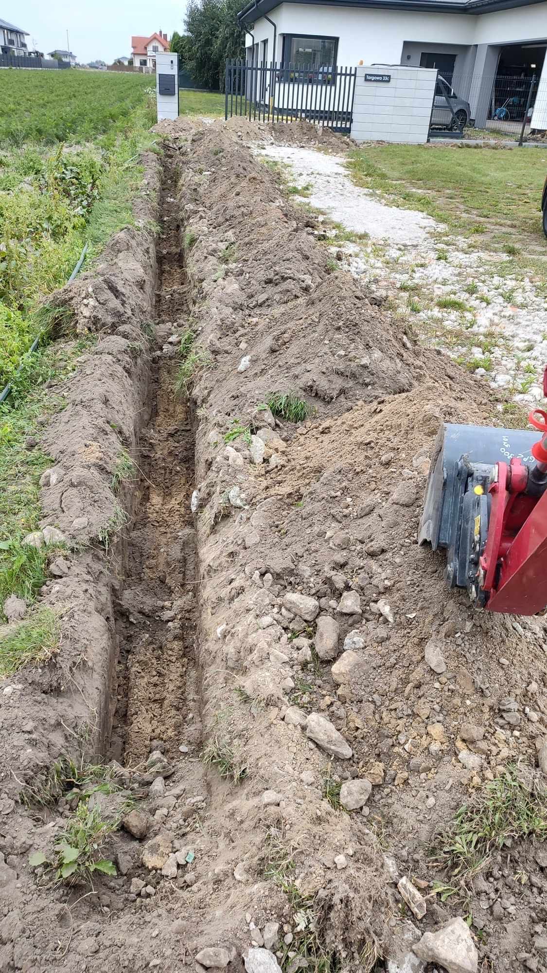 Długi, wąski wykop w ziemi, wykonany blisko domu jednorodzinnego, z widocznym fragmentem koparki z boku.