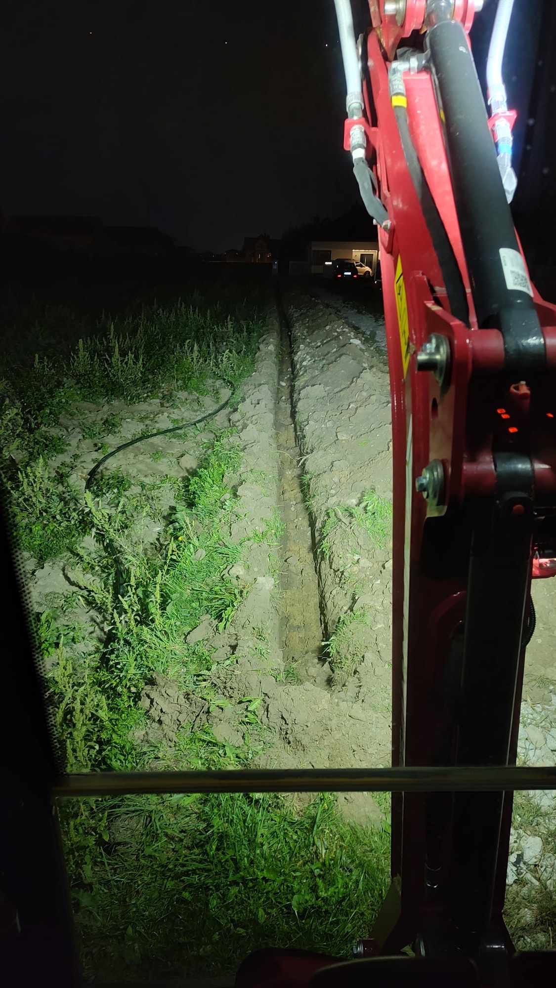 Wykopany rów w ziemi widziany z kabiny czerwonej koparki w nocy, oświetlony światłami maszyny, z widoczną rurą wzdłuż rowu i zabudowaniami w tle.