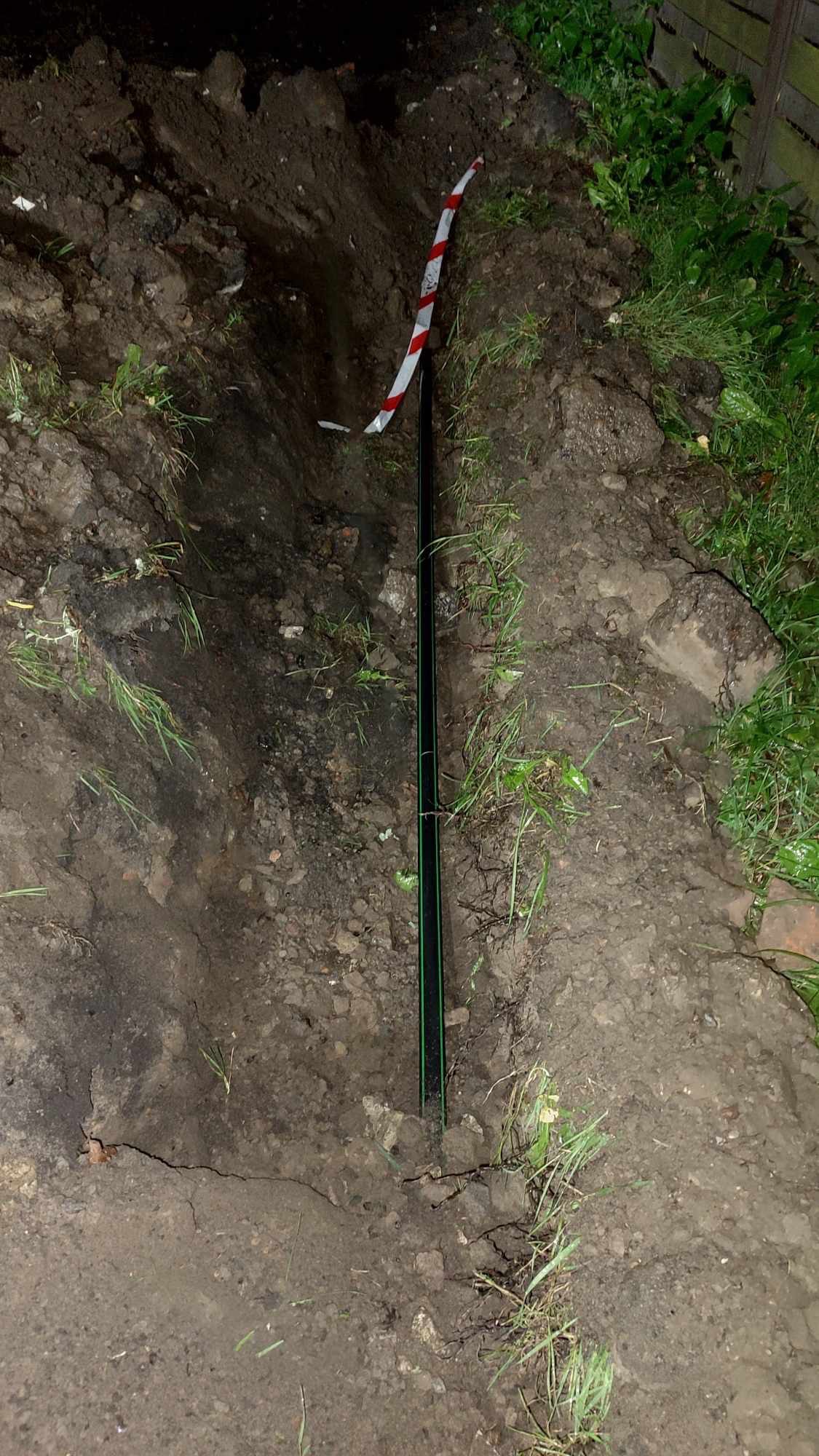 Wykop w ziemi z widoczną rurą w kolorze czarnym z zielonym paskiem, częściowo zasypany ziemią, z taśmą ostrzegawczą biało-czerwoną w tle.