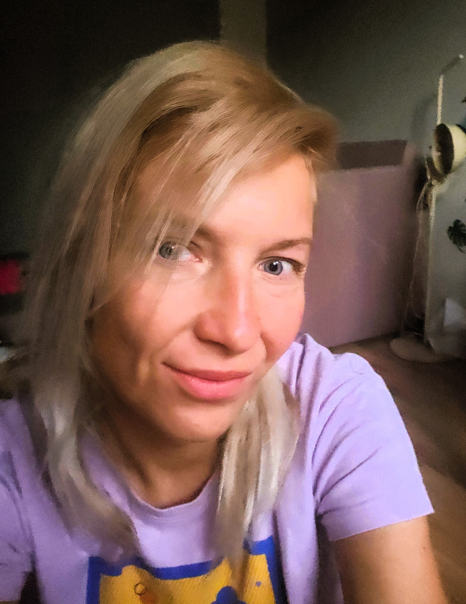 Portret uśmiechniętej blondynki w fioletowej koszulce, patrzącej w obiektyw, z lekkim rozmyciem tła.