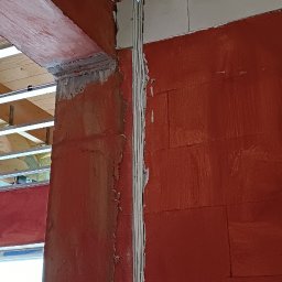 ALTROM Łukasz Olesiak - Wykonane bruzdy w czerwonej ścianie z betonu komórkowego, w których ułożono białe przewody elektryczne, widoczne metalowe profile pod sufitem.