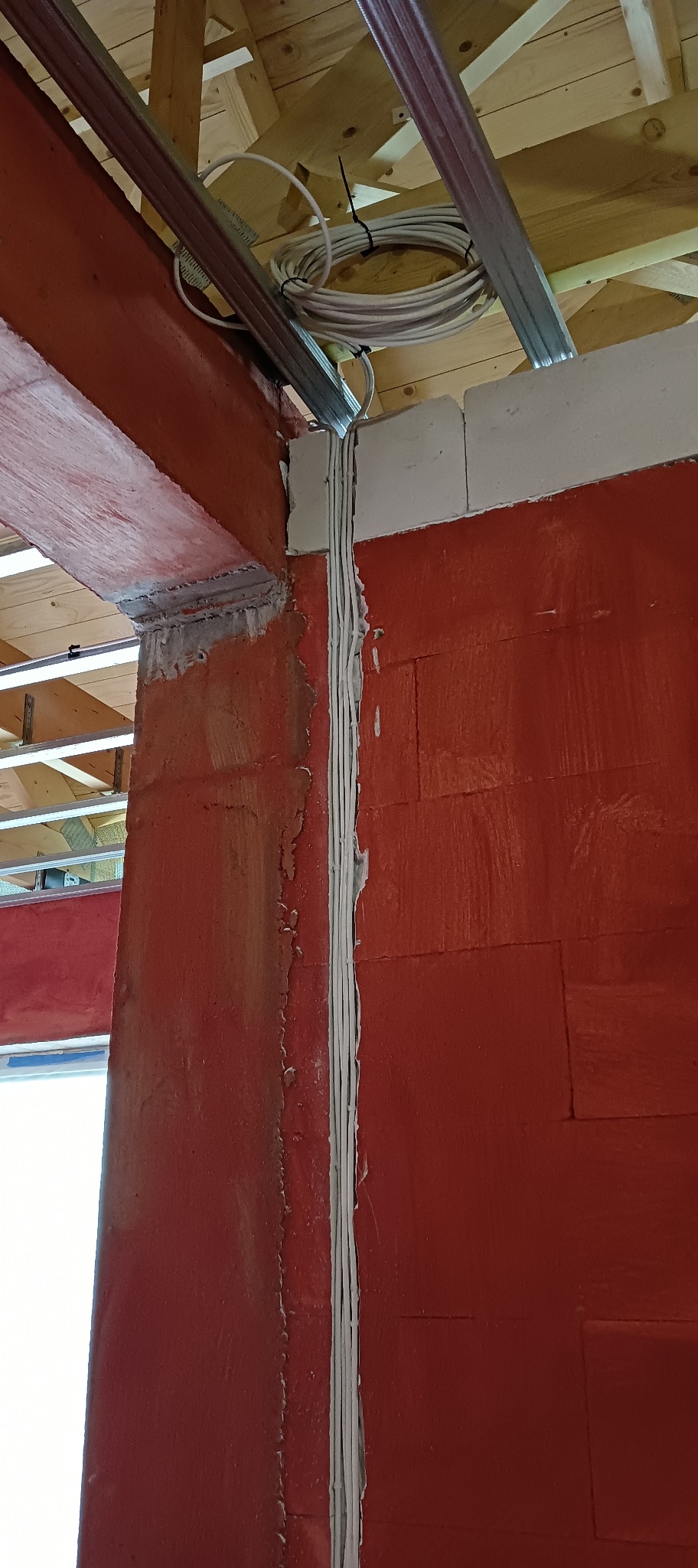 Wykonane bruzdy w czerwonej ścianie z betonu komórkowego, w których ułożono białe przewody elektryczne, widoczne metalowe profile pod sufitem.