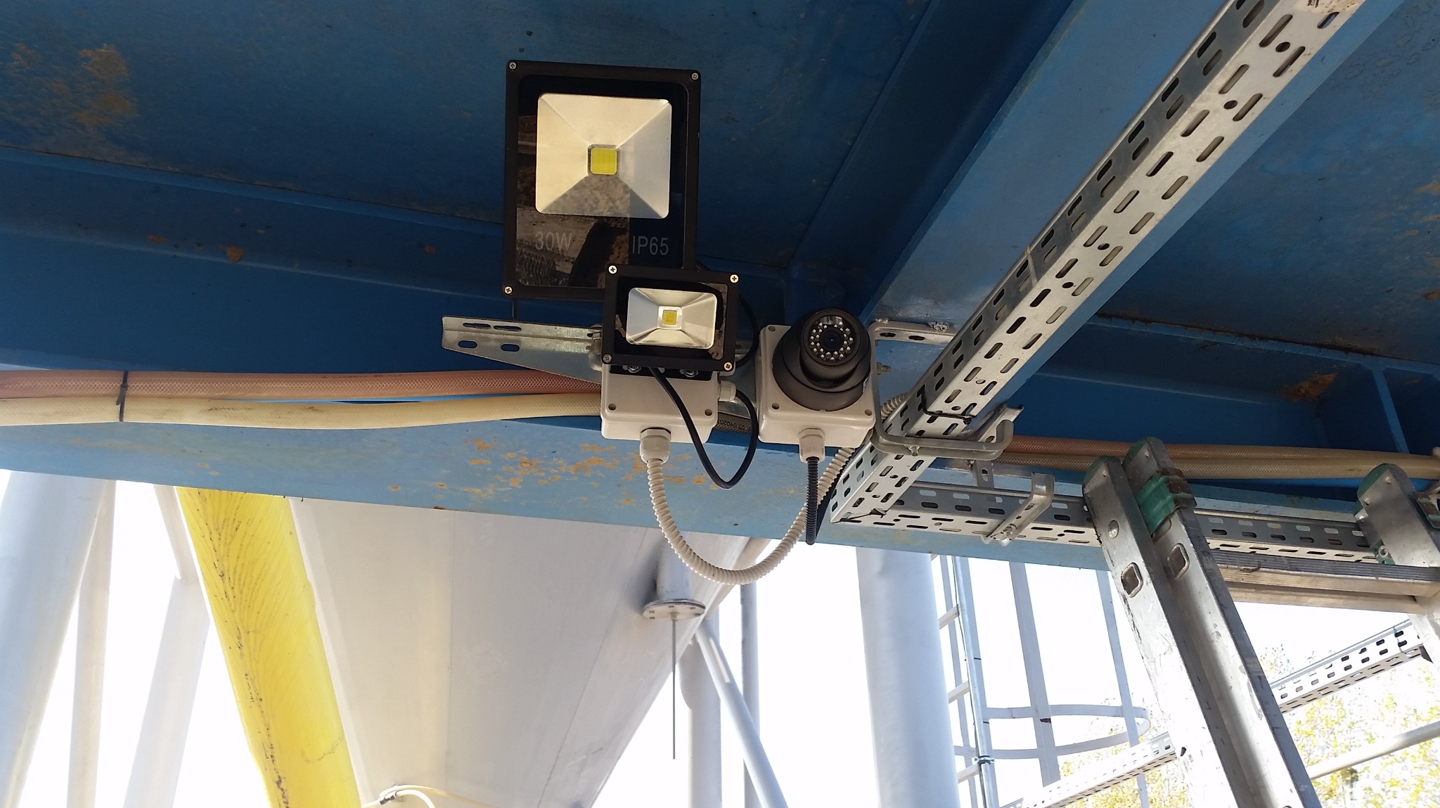 Kamera monitoringu kopułkowa i dwa reflektory LED (30W i mniejszy) zamontowane na metalowej konstrukcji pod sufitem, widoczne kable w peszlach i drabina aluminiowa w tle.