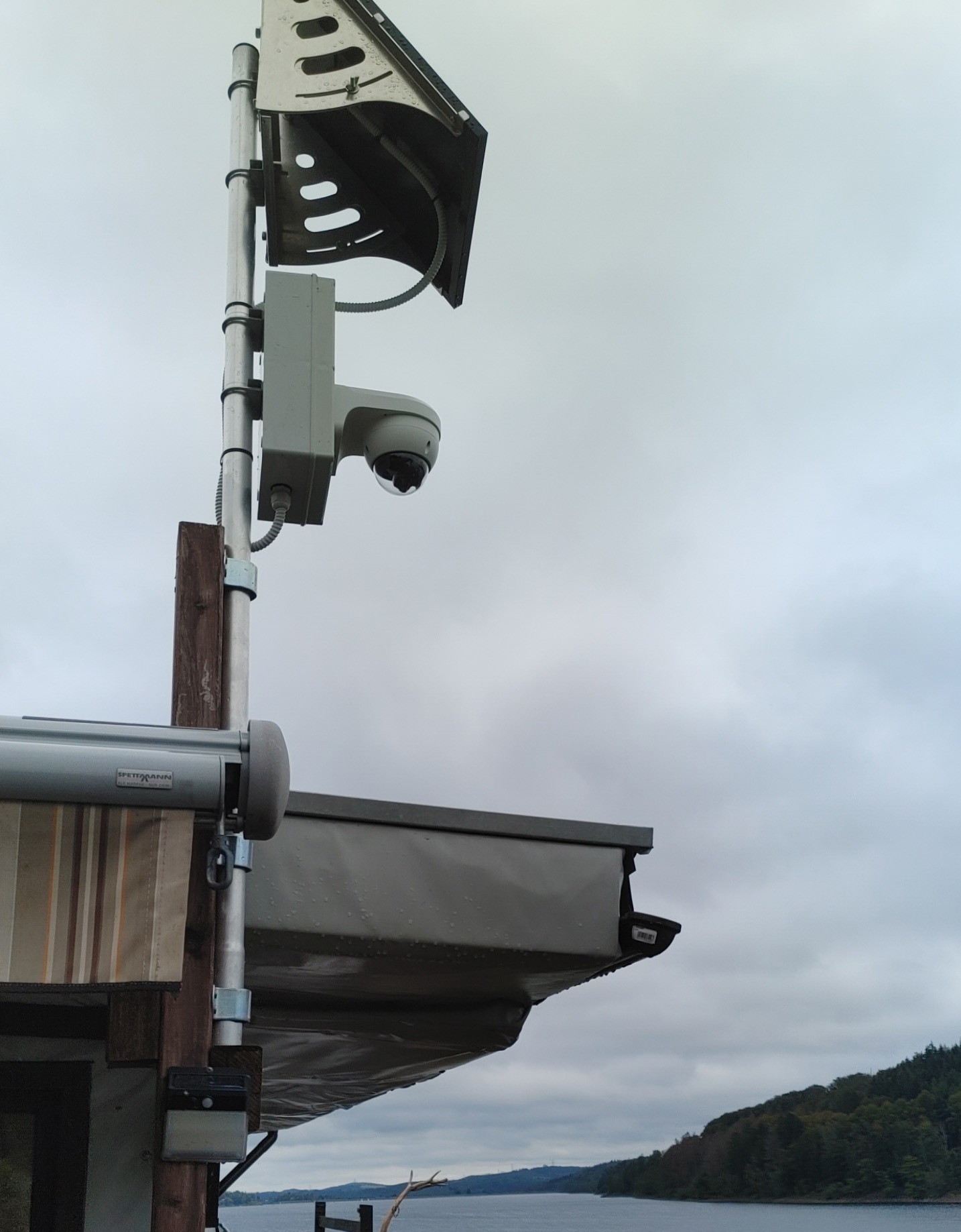 Zewnętrzna kamera monitoringu kopułkowa zamontowana na metalowym słupku z osłoną przeciwsłoneczną, widok na jezioro i zalesiony brzeg w tle, pochmurne niebo.