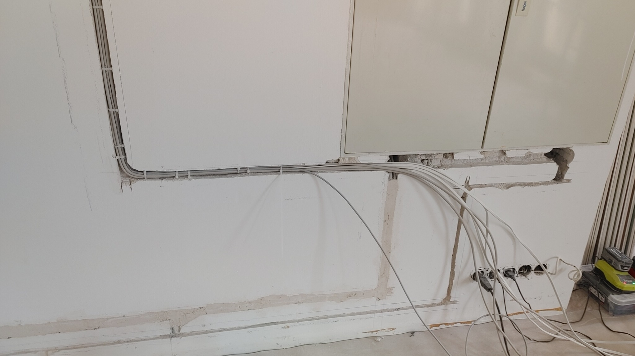 Biała ściana z wybruzdowanymi kanałami, w których ułożone są szare przewody elektryczne, częściowo przymocowane uchwytami, z puszkami elektrycznymi i narzędziami Ryobi w tle.
