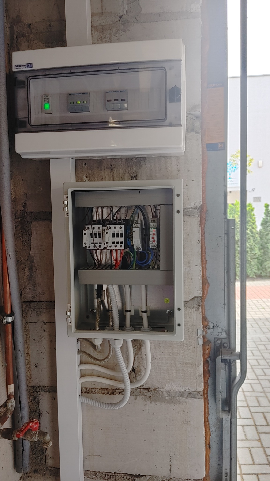 Otwarta skrzynka elektryczna z widocznymi bezpiecznikami, przekaźnikami i przewodami na szarej ścianie z betonu komórkowego, obok rury i drzwi garażowych.