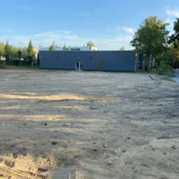 Przygotowany teren pod budowę, z widocznymi śladami maszyn i niewielkim drzewem przed szarym budynkiem z białymi drzwiami.