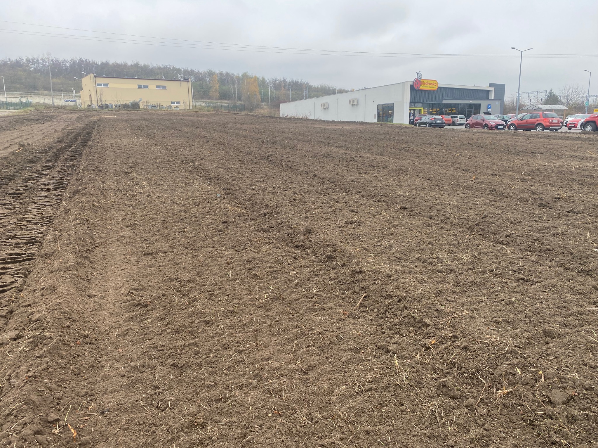 Przygotowany teren pod budowę, świeżo wyrównana ziemia z widocznymi śladami opon, w tle budynki handlowe i magazynowe w pochmurny dzień.