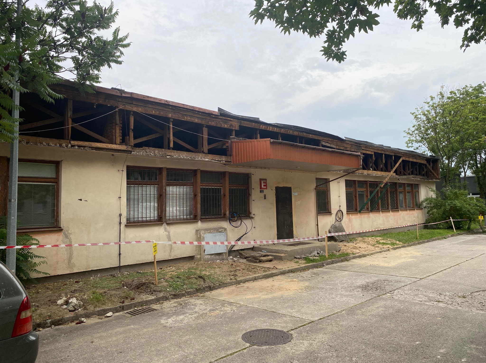 Częściowo zdemontowany budynek z odsłoniętą konstrukcją dachu, zabezpieczony taśmą ostrzegawczą, z widocznymi śladami gruzu i rozbiórki.