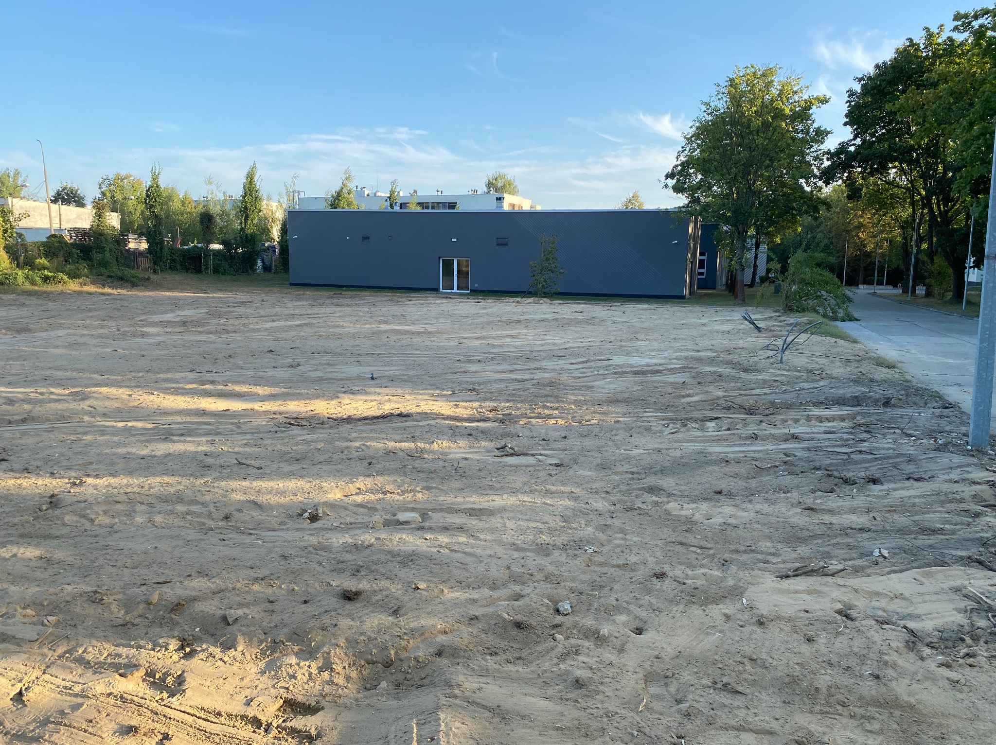 Przygotowany teren pod budowę, z widocznymi śladami maszyn i niewielkim drzewem przed szarym budynkiem z białymi drzwiami.