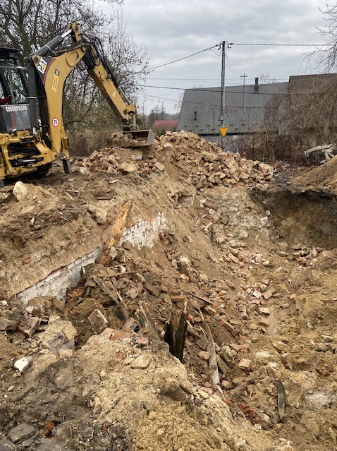Żółta koparka na placu budowy, gruzowisko po rozbiórce budynku z fragmentami cegieł i drewna, widoczne fundamenty. Prace ziemne w toku.