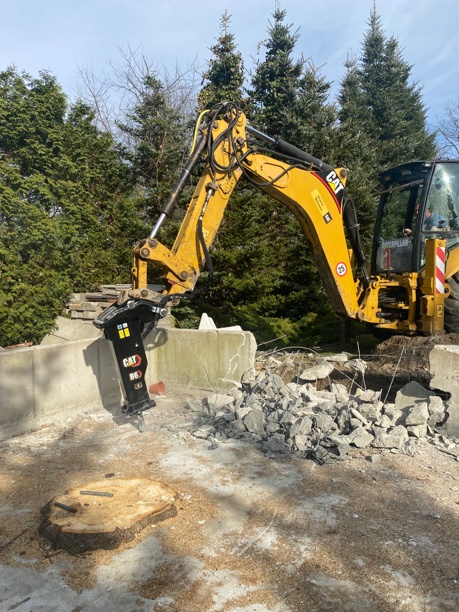 Żółta koparka CAT z hydraulicznym młotem pneumatycznym rozbija betonową konstrukcję. W tle drzewa i niebieskie niebo. Widoczny pień drzewa.