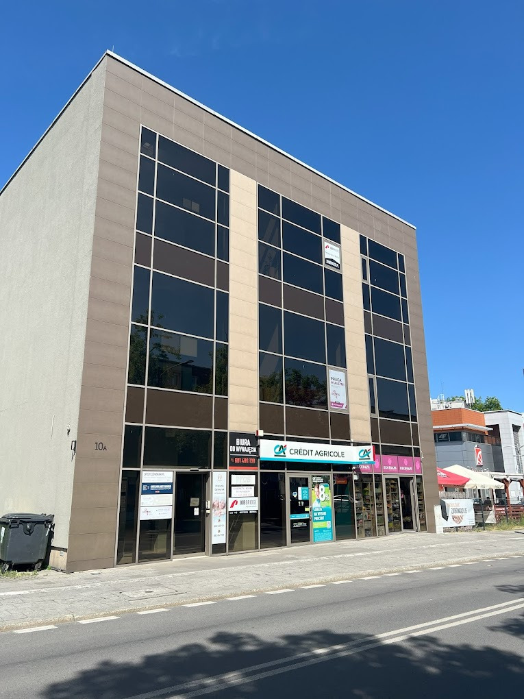 Elewacja biurowca z brązowymi panelami i ciemnymi oknami, z widocznymi szyldami firm na parterze, w tym Credit Agricole, oraz ogłoszeniami o wynajmie biur.