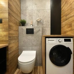 Taupe Interior - Nowoczesna łazienka z drewnianymi panelami ściennymi, szarą płytką imitującą beton, podwieszaną toaletą i pralką zabudowaną w drewnianej obudowie.
