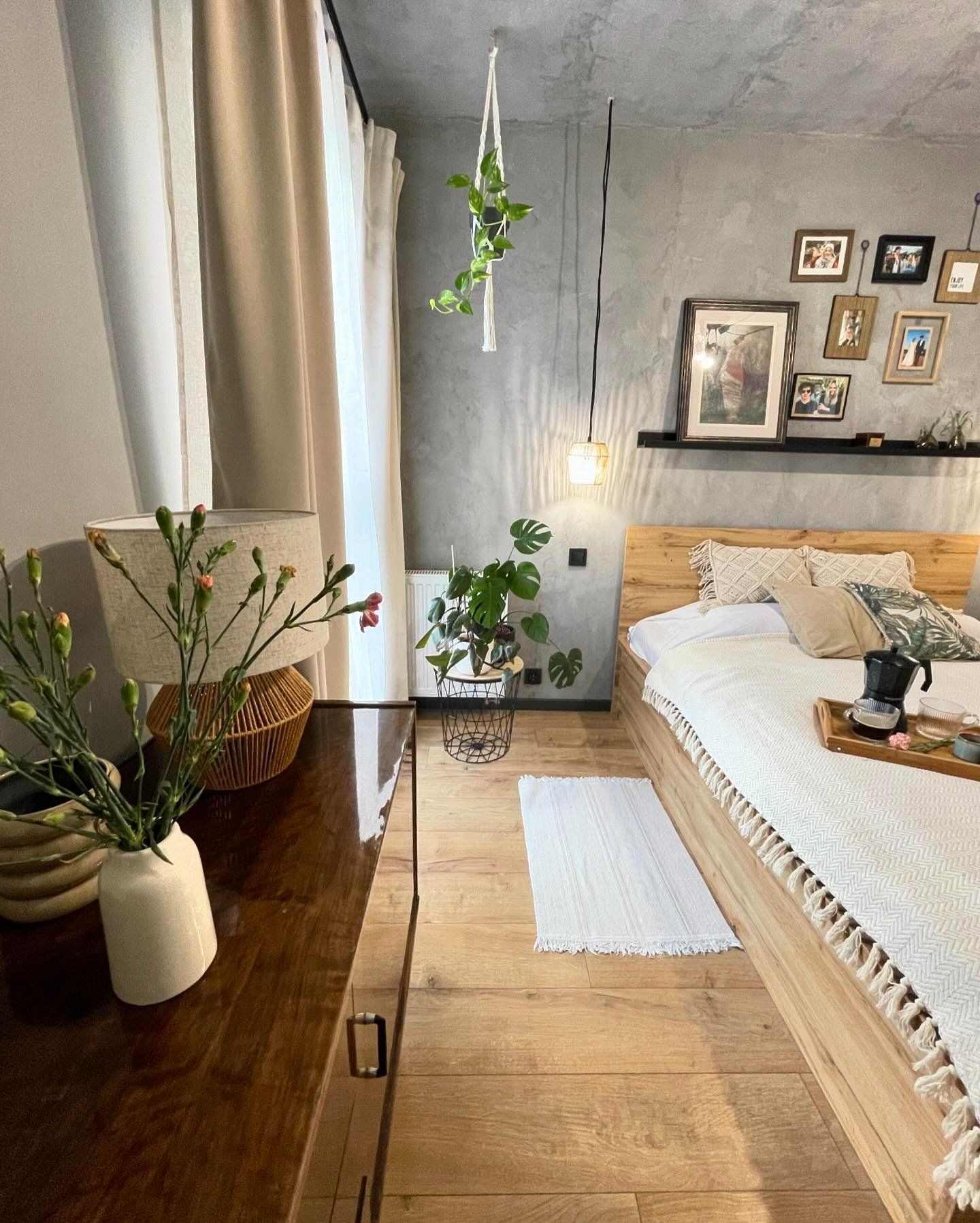 Sypialnia w stylu boho z elementami industrialnymi: drewniane łóżko z białą narzutą w jodełkę i frędzlami, stolik nocny z wazonem i lampką, szara ściana z galerią zdjęć i wiszącą rośliną,...