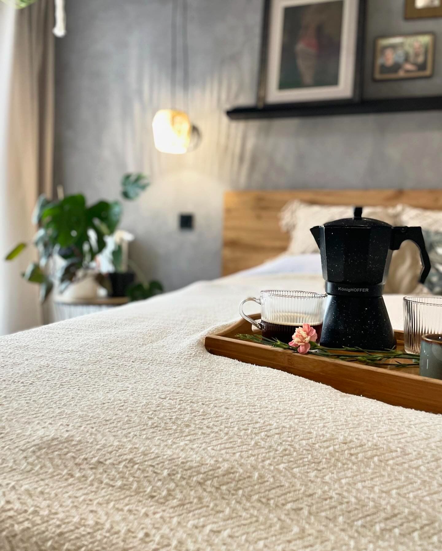 Śniadanie na drewnianej tacy w sypialni: czarna kafeterka KönigHOFFER, szklanka z kawą, szklanka z wodą, mały różowy kwiat i gałązki na tle łóżka z beżową narzutą i betonowej ściany z obrazami.