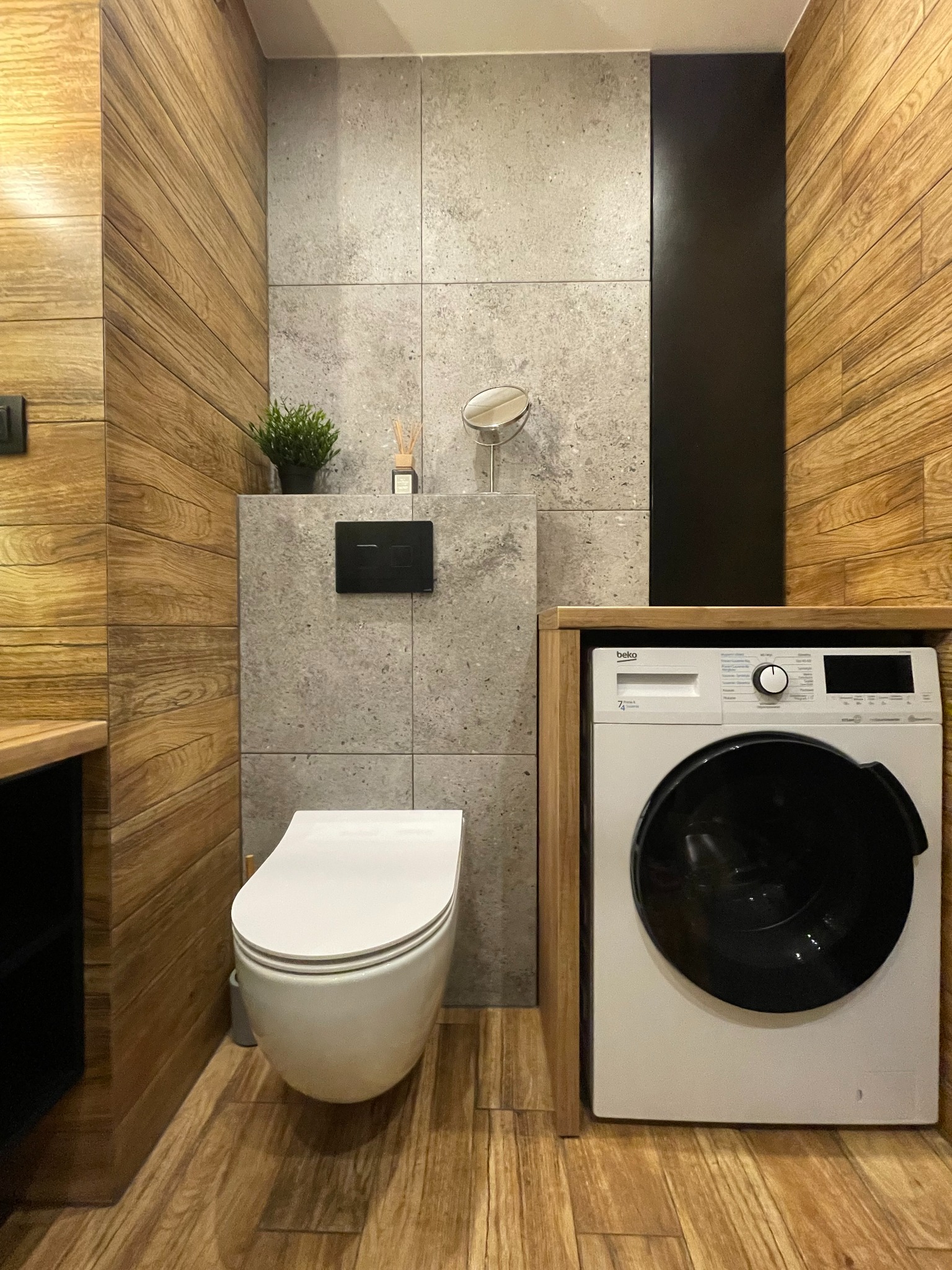 Nowoczesna łazienka z drewnianymi panelami ściennymi, szarą płytką imitującą beton, podwieszaną toaletą i pralką zabudowaną w drewnianej obudowie.
