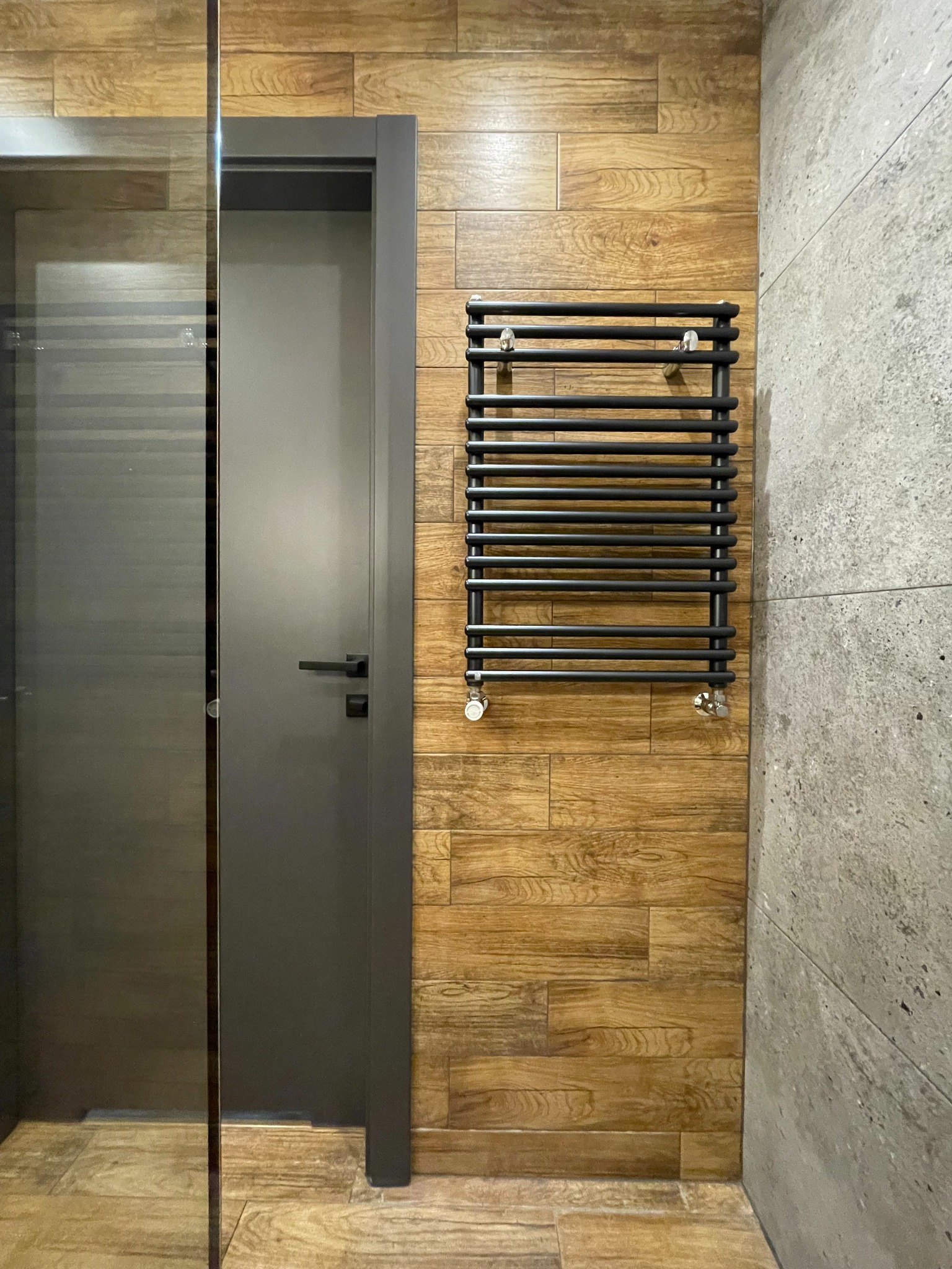 Nowoczesna łazienka z drewnopodobnymi płytkami na ścianie, grafitową drabinką grzejnikową i szarą ścianą z betonu architektonicznego, widoczna szklana kabina prysznicowa.