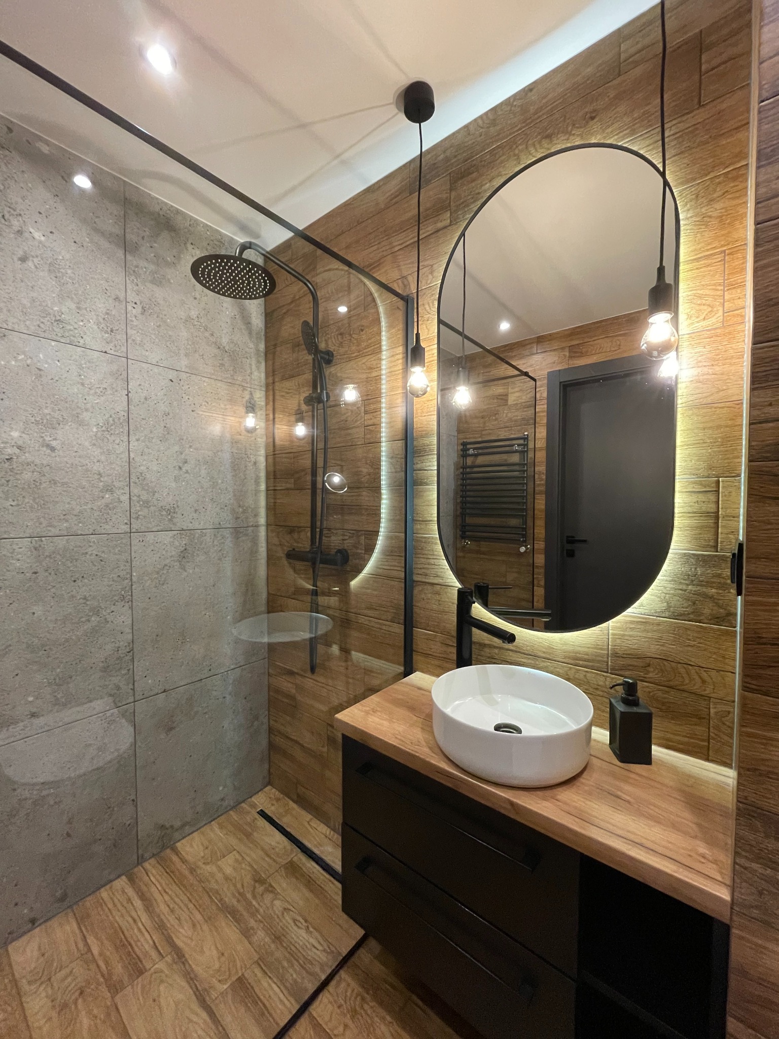 Nowoczesna łazienka w stylu industrialnym z drewnianymi akcentami: kabina prysznicowa typu walk-in z deszczownicą, owalne lustro z podświetleniem LED, biała umywalka nablatowa na drewnianym blacie...