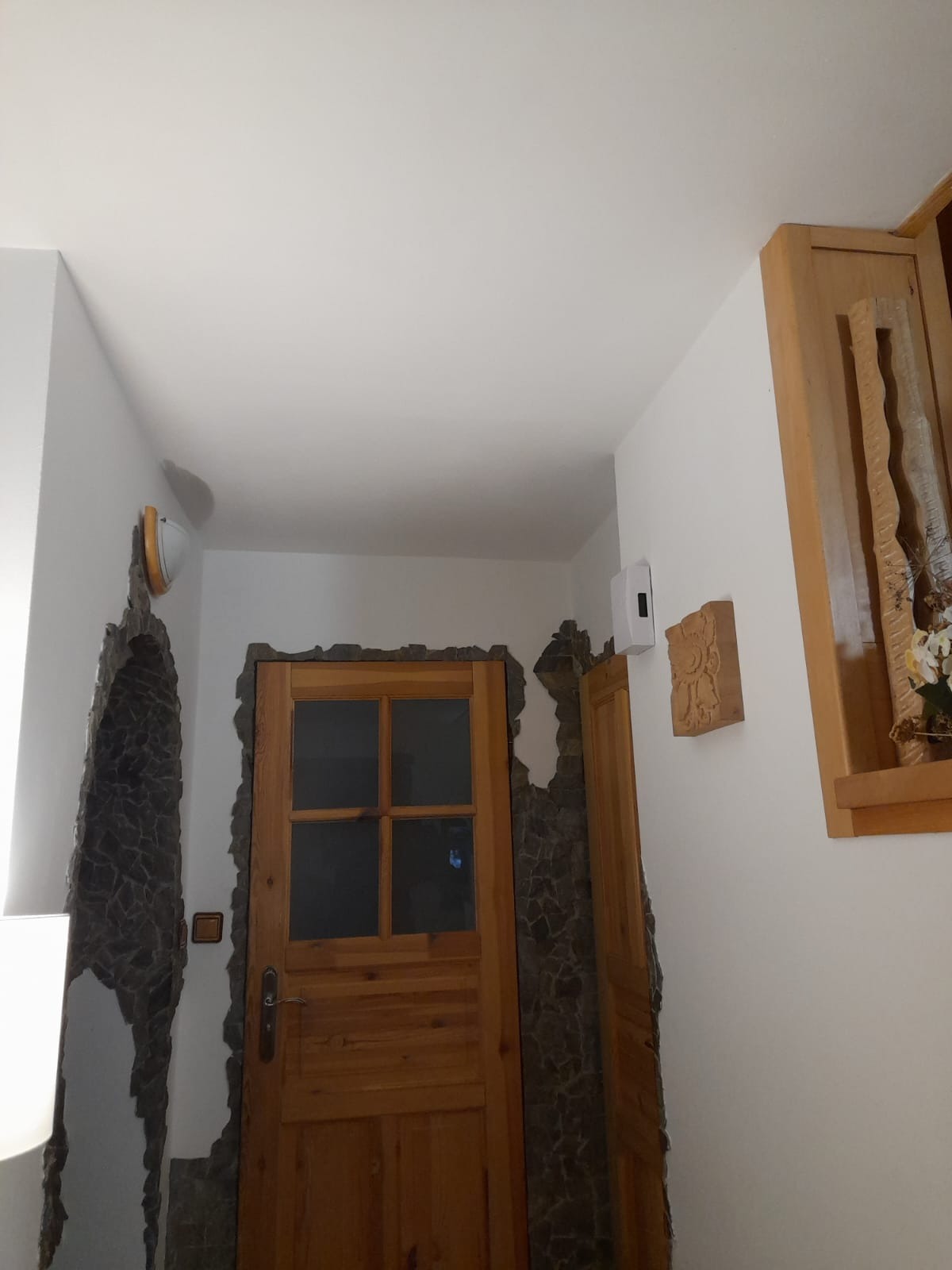 Drewniane drzwi z szybami, otoczone dekoracyjnym kamieniem na ścianie, z widoczną drewnianą płaskorzeźbą i półką z dekoracją kwiatową w tle.