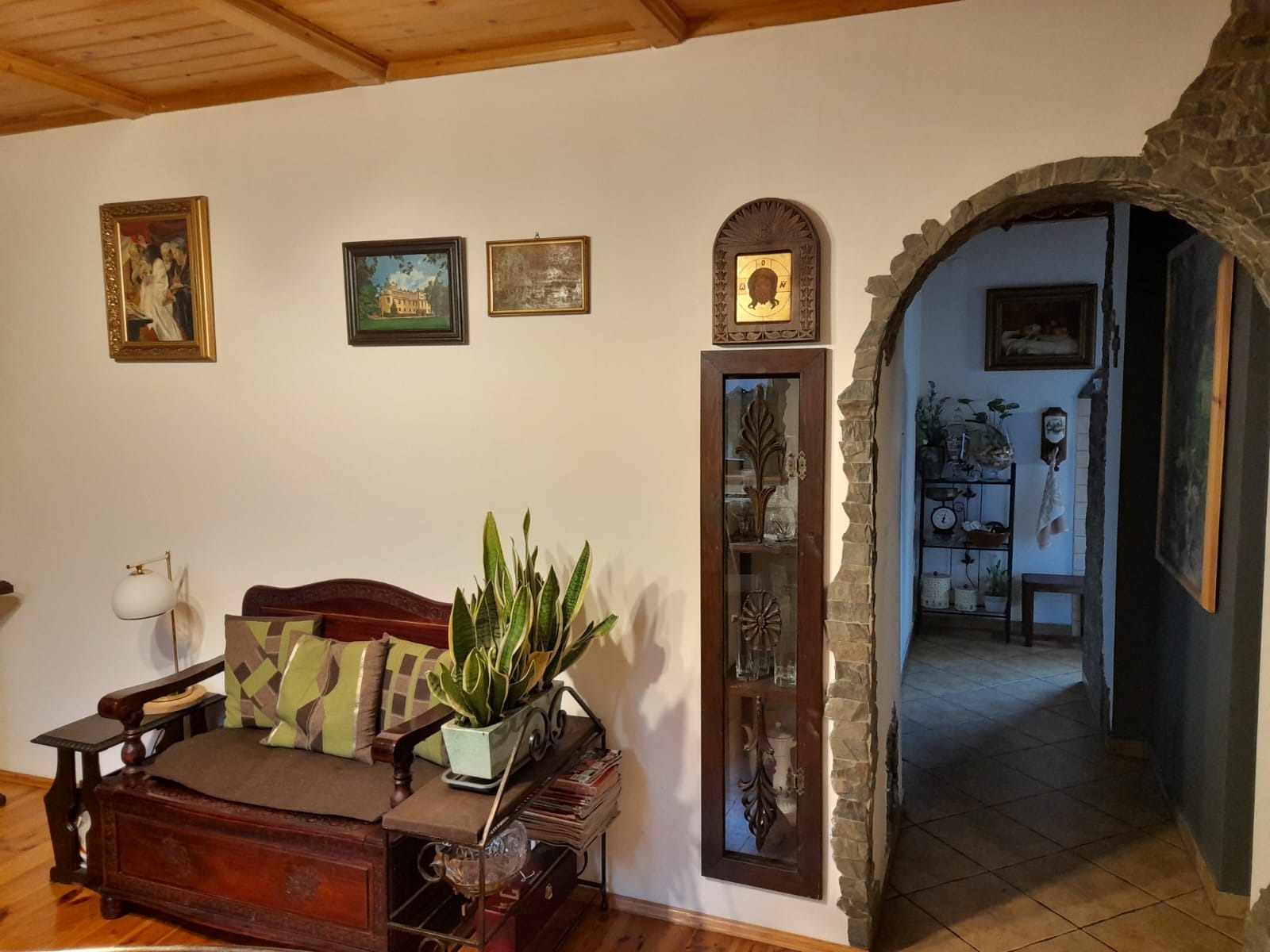 Rustykalny salon z drewnianą ławą skrzyniową, zielonymi poduszkami, rośliną doniczkową i kamiennym łukiem prowadzącym do dalszej części domu.