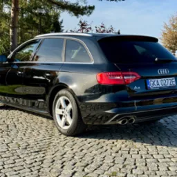 Czarny samochód marki Audi A4 Avant zaparkowany na brukowanej nawierzchni, widok z tyłu i boku, z widocznymi detalami jak tablica rejestracyjna, felgi i podwójny wydech.