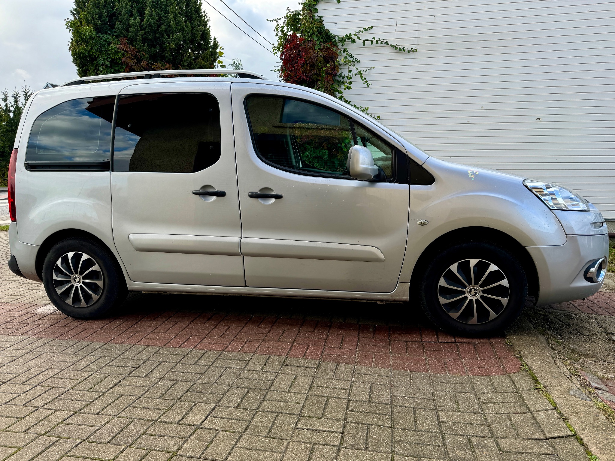 Srebrny van zaparkowany na brukowanej nawierzchni przed białym budynkiem z pnącą roślinnością, widok z boku.