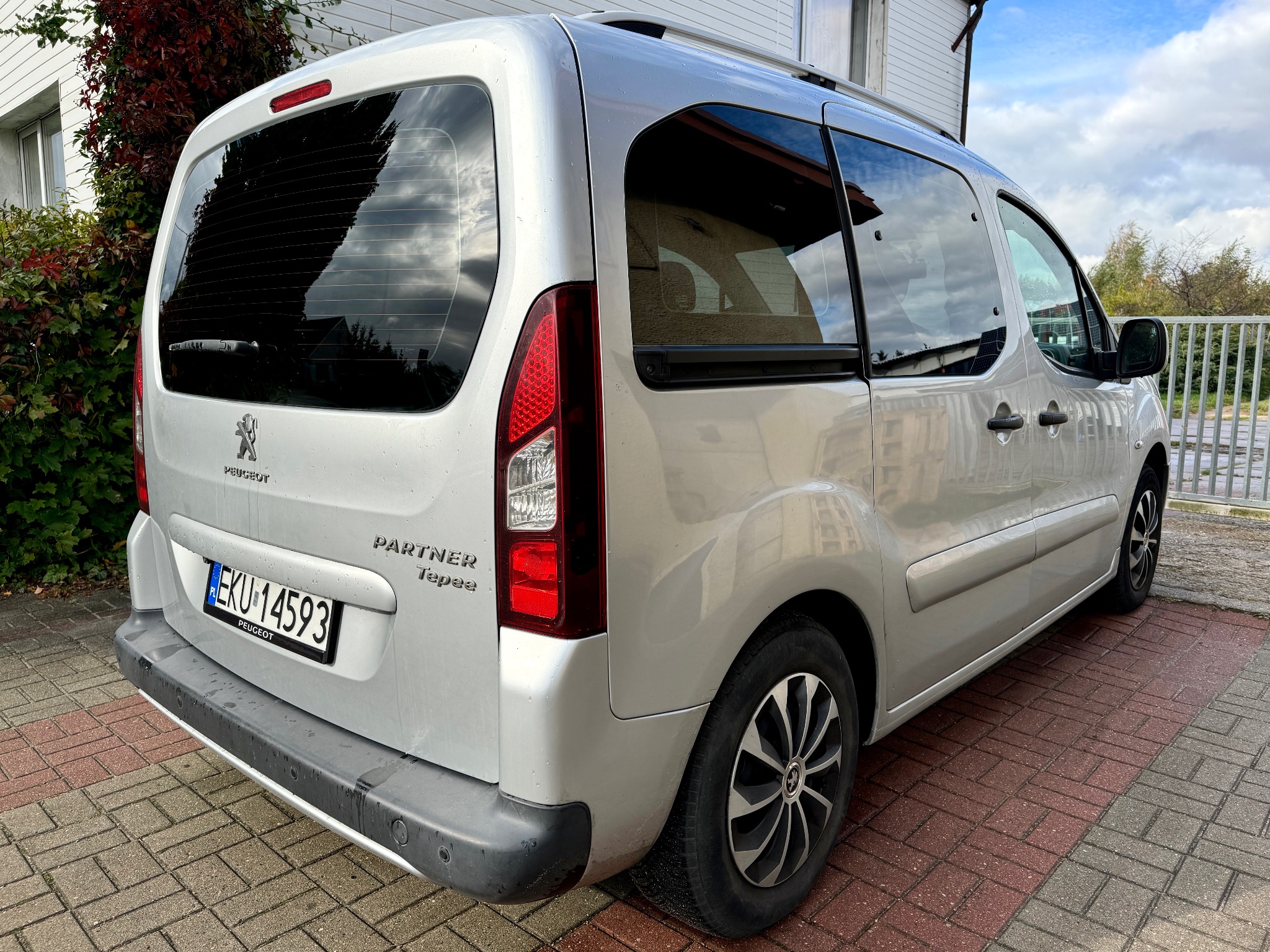 Srebrny Peugeot Partner Tepee zaparkowany na kostce brukowej, widok z tyłu, z widoczną tablicą rejestracyjną EKU-14593, z elementami roślinności w tle.
