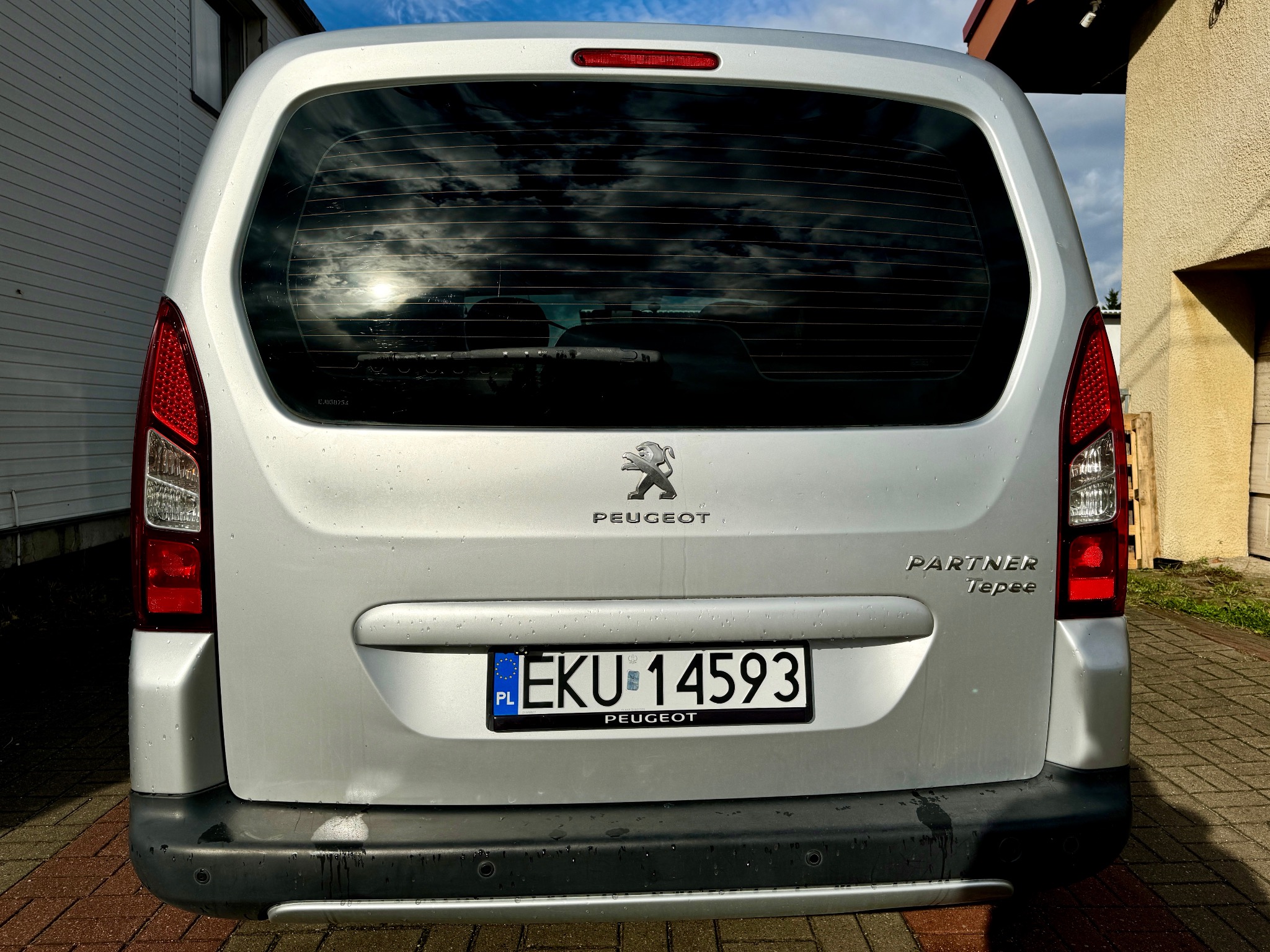 Tył srebrnego samochodu Peugeot Partner Tepee z polską tablicą rejestracyjną EKU 14593, widoczne krople wody na karoserii i odbicie chmur w tylnej szybie.