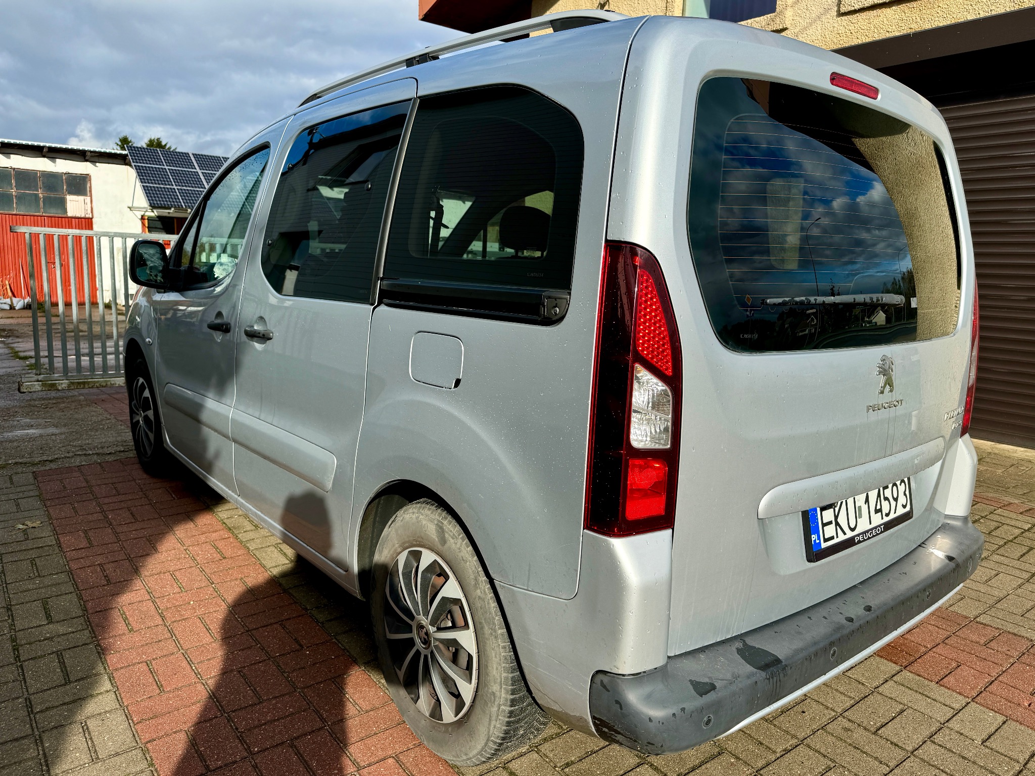 Srebrny Peugeot Partner z polską tablicą rejestracyjną EKU 14593, zaparkowany na czerwonej kostce brukowej przed budynkiem z panelami słonecznymi.