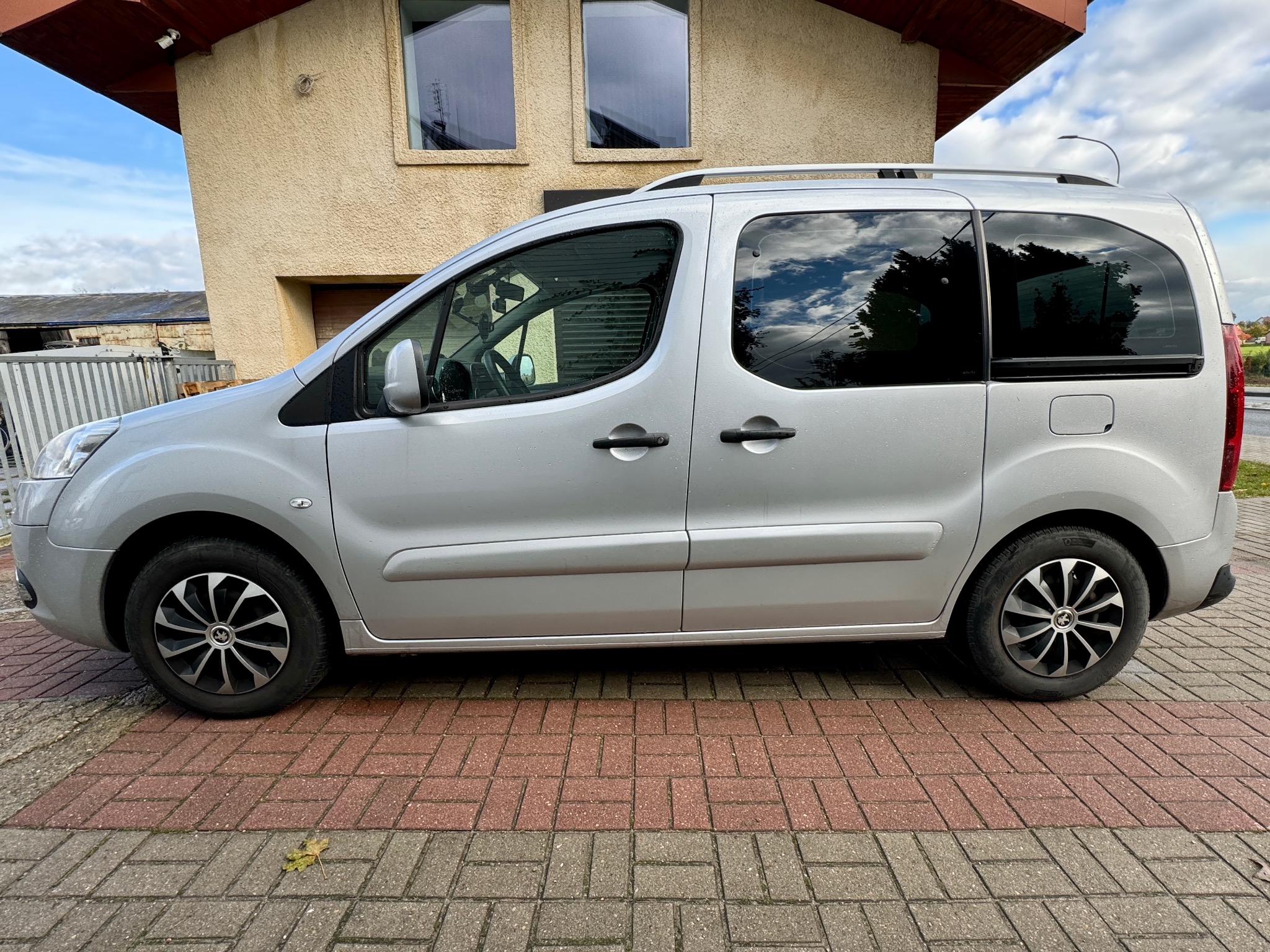 Srebrny samochód typu kombivan zaparkowany na brukowanej nawierzchni przed budynkiem z beżową elewacją, widoczne odbicie chmur na przyciemnianych szybach.