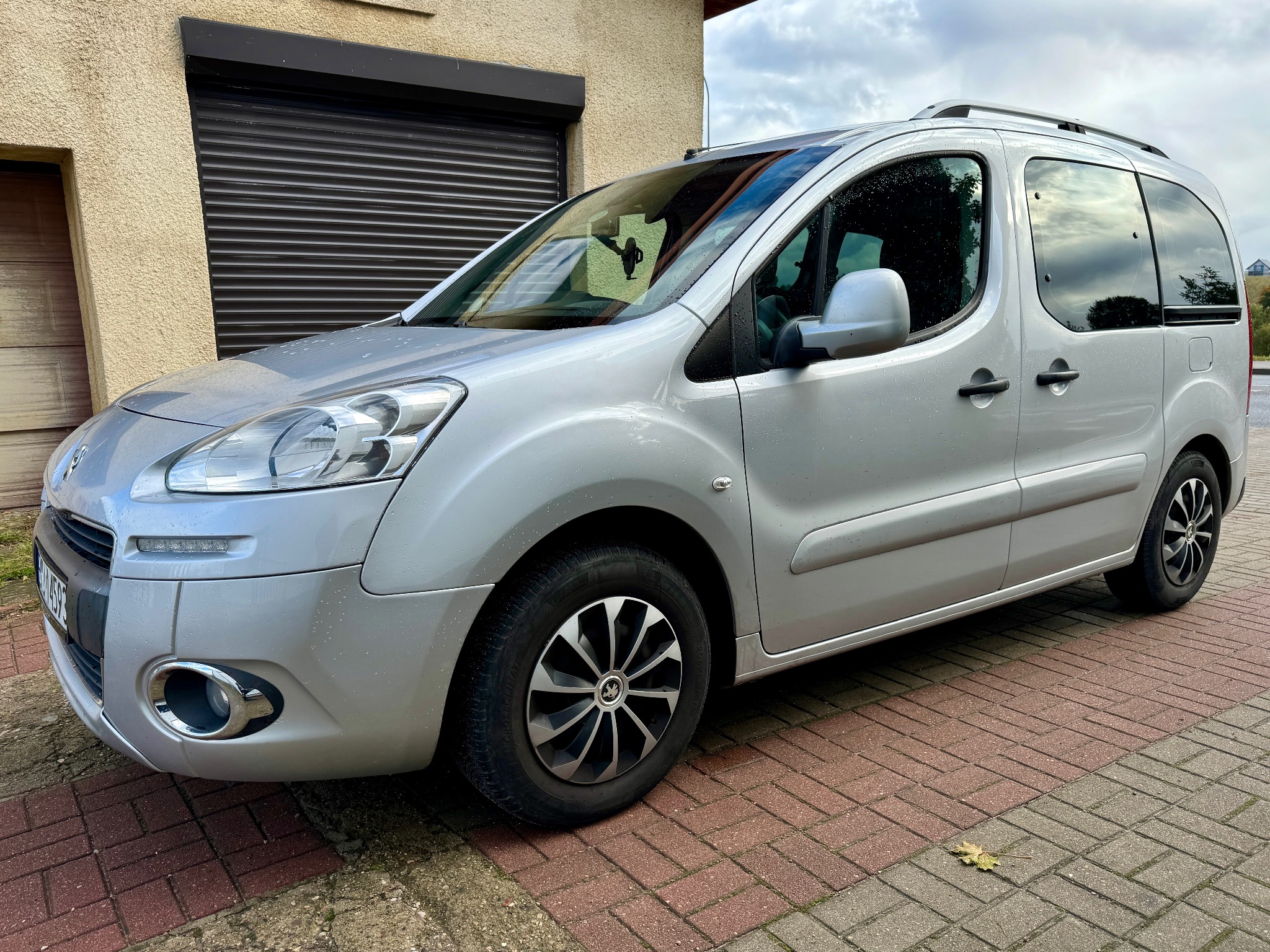 Srebrny samochód typu kombivan zaparkowany na kostce brukowej przed budynkiem z brązową bramą roletową.