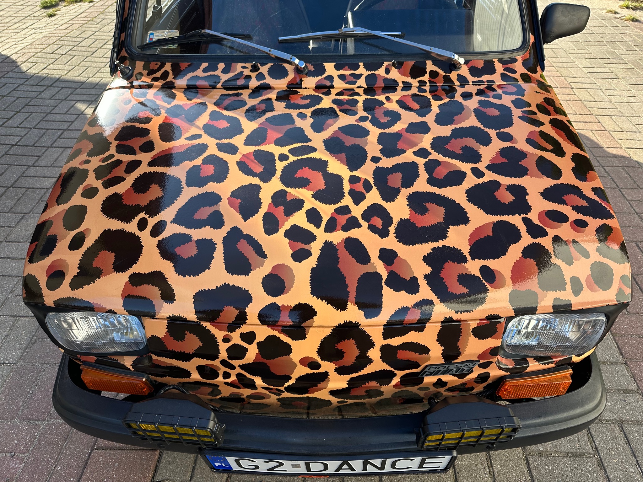 Mały Fiat 126p z oklejeniem w panterkę, widoczne przednie światła i tablica rejestracyjna G2-DANCE.
