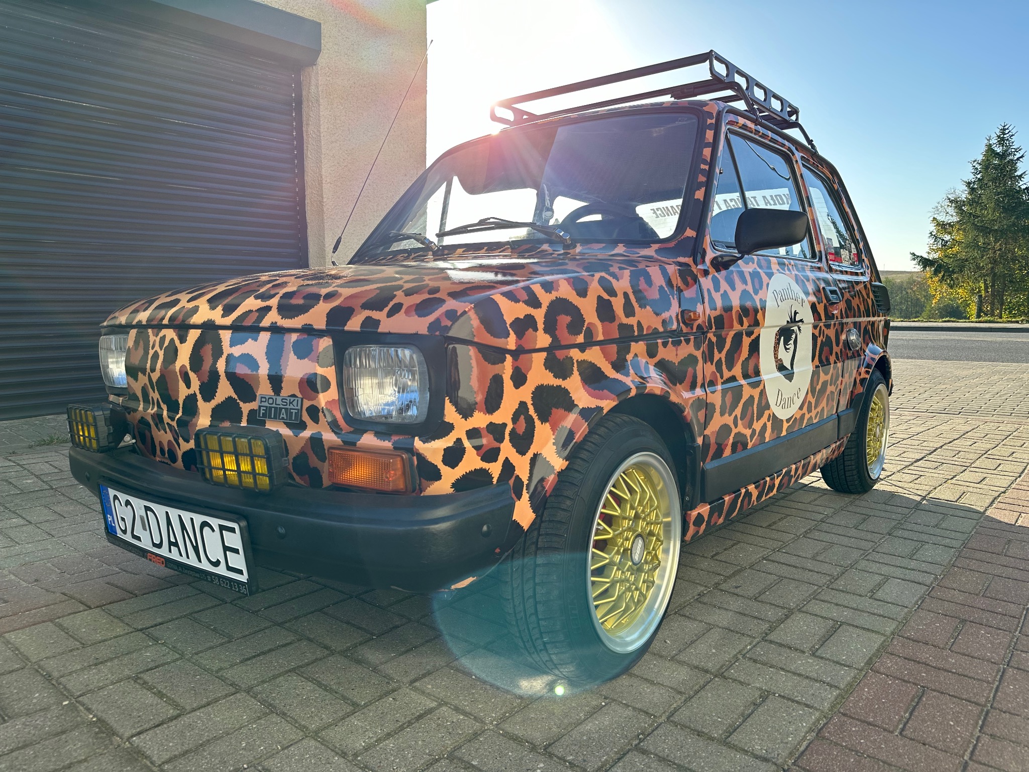 Maluch w panterkę z logo 'Panther Dance' na boku, z bagażnikiem dachowym i złotymi felgami, zaparkowany na kostce brukowej.