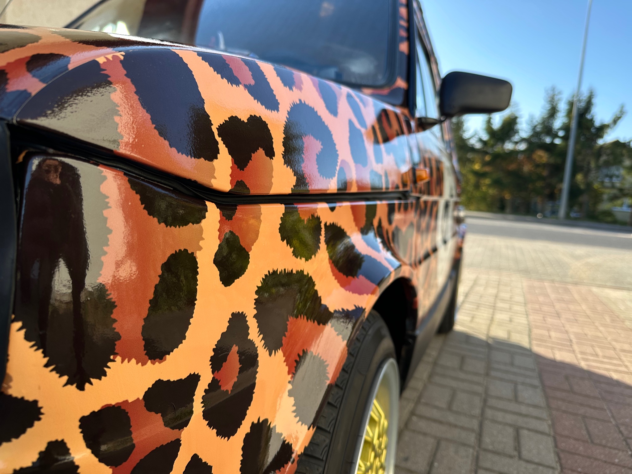 Samochód Fiat 126p oklejony folią w panterkę, z widocznymi złotymi felgami, zaparkowany na kostce brukowej w słoneczny dzień.
