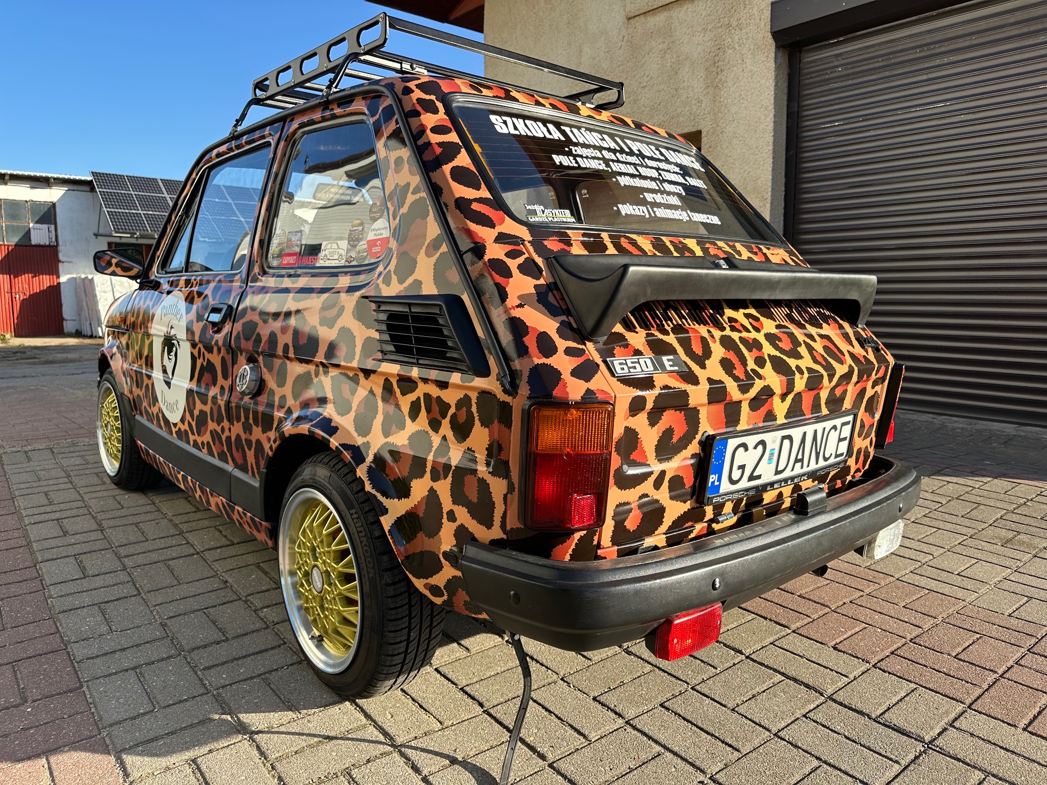 Samochód Fiat 126p oklejony folią w panterkę z napisem 'Szkoła Tańca i Pole Dance' na tylnej szybie, na złotych felgach, z bagażnikiem dachowym
