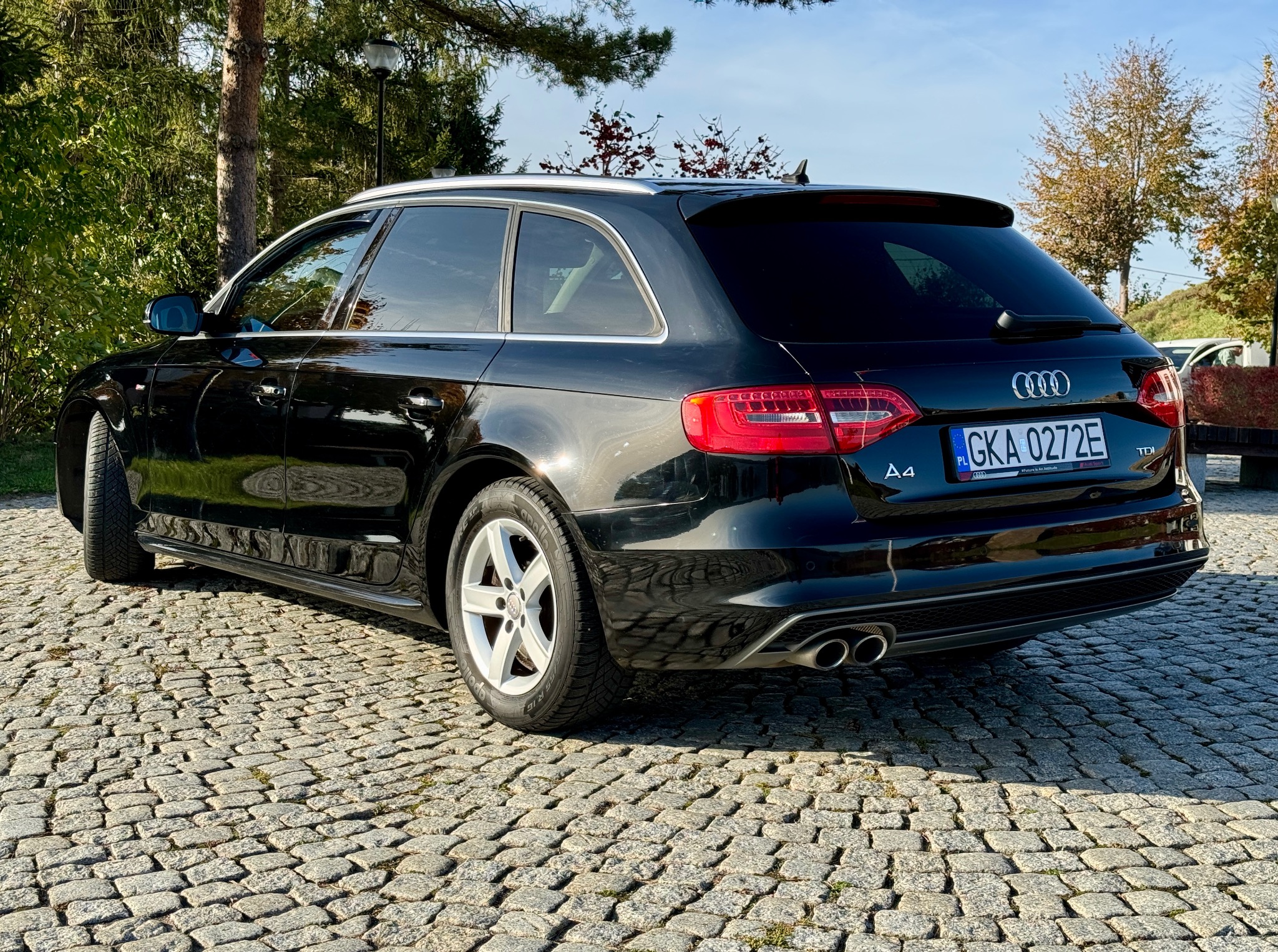 Czarny samochód marki Audi A4 Avant zaparkowany na brukowanej nawierzchni, widok z tyłu i boku, z widocznymi detalami jak tablica rejestracyjna, felgi i podwójny wydech.