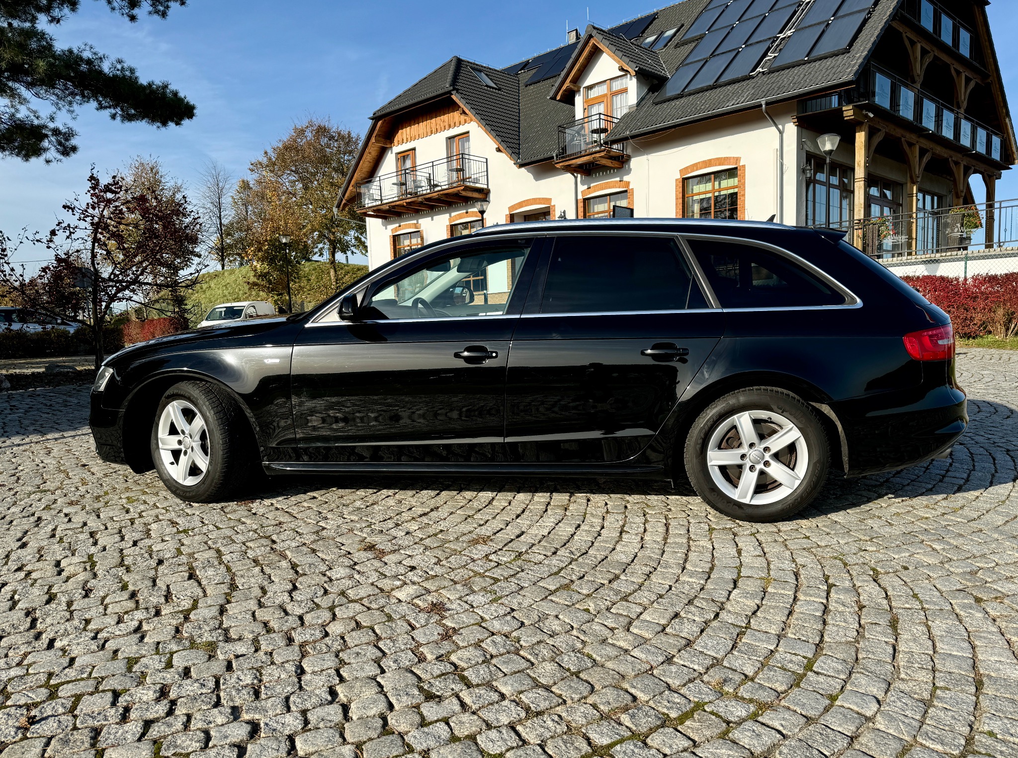 Czarny samochód kombi zaparkowany na brukowanej nawierzchni przed domem z panelami słonecznymi na dachu.
